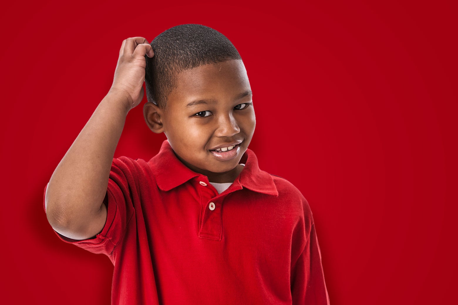 A little boy wearing a red polo shirt scratches his head and looks bewildered.