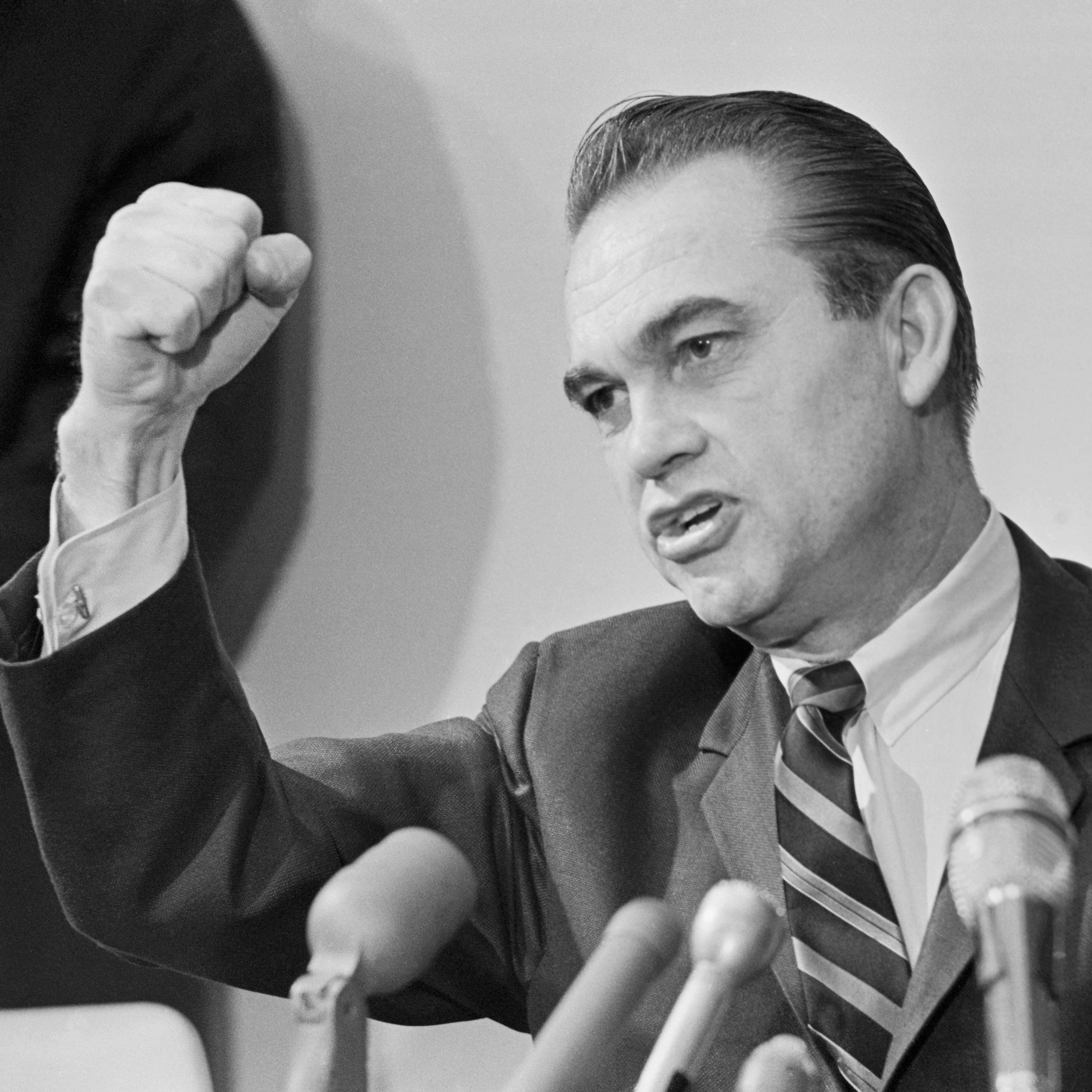 Wallace speaking seated in front of microphones with his right fist raised in the air