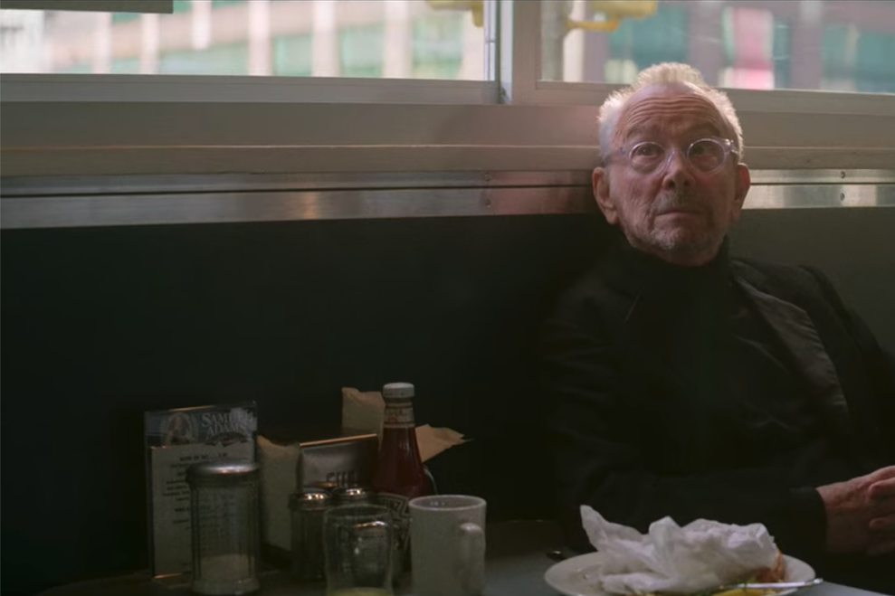 Joel Grey sits with his hands clasped together at a diner table.