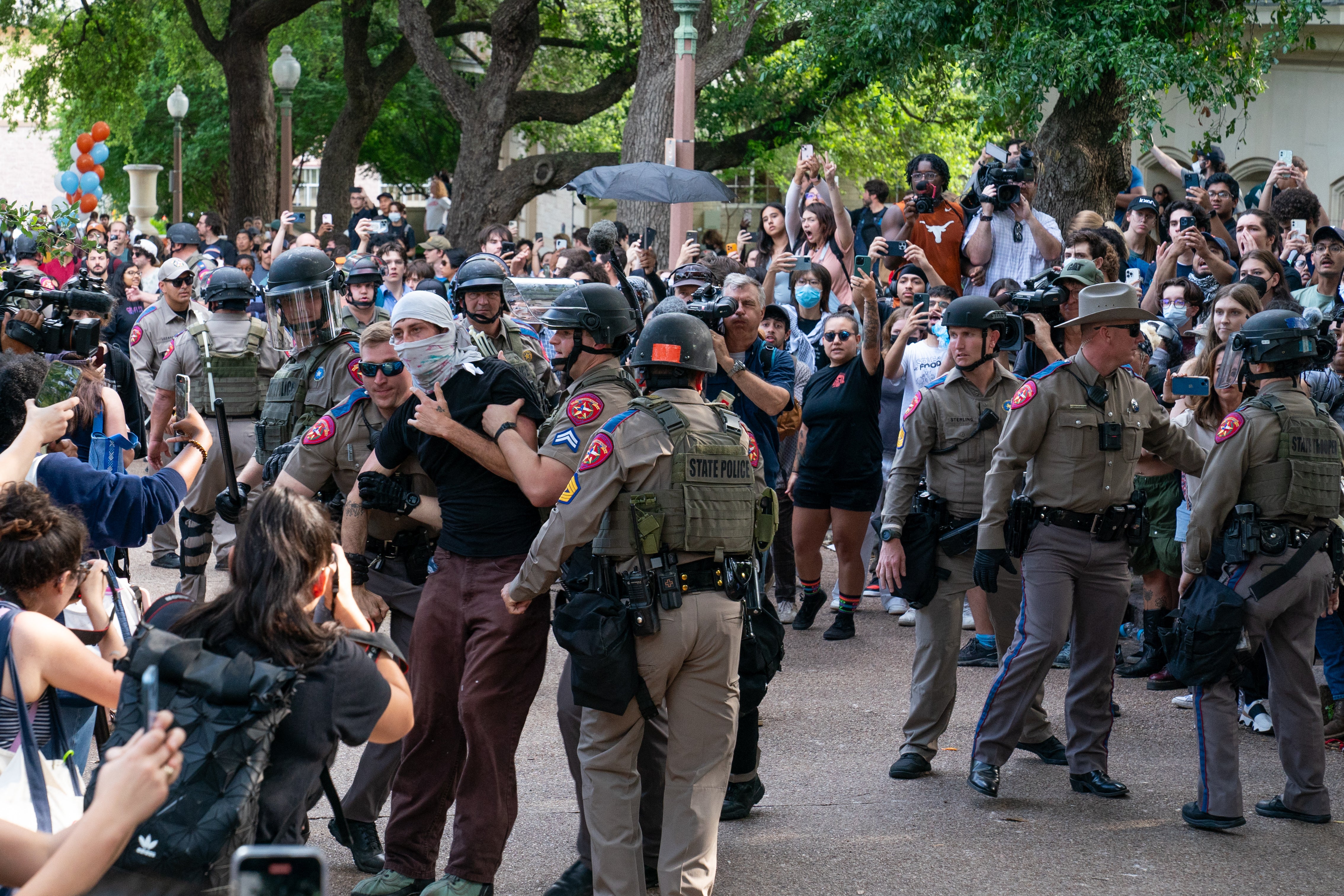 State troopers walk into a crowd of student protesters on a campus; most students are taking pictures; 3 officers are restraining one protester by both arms as other officers look on.