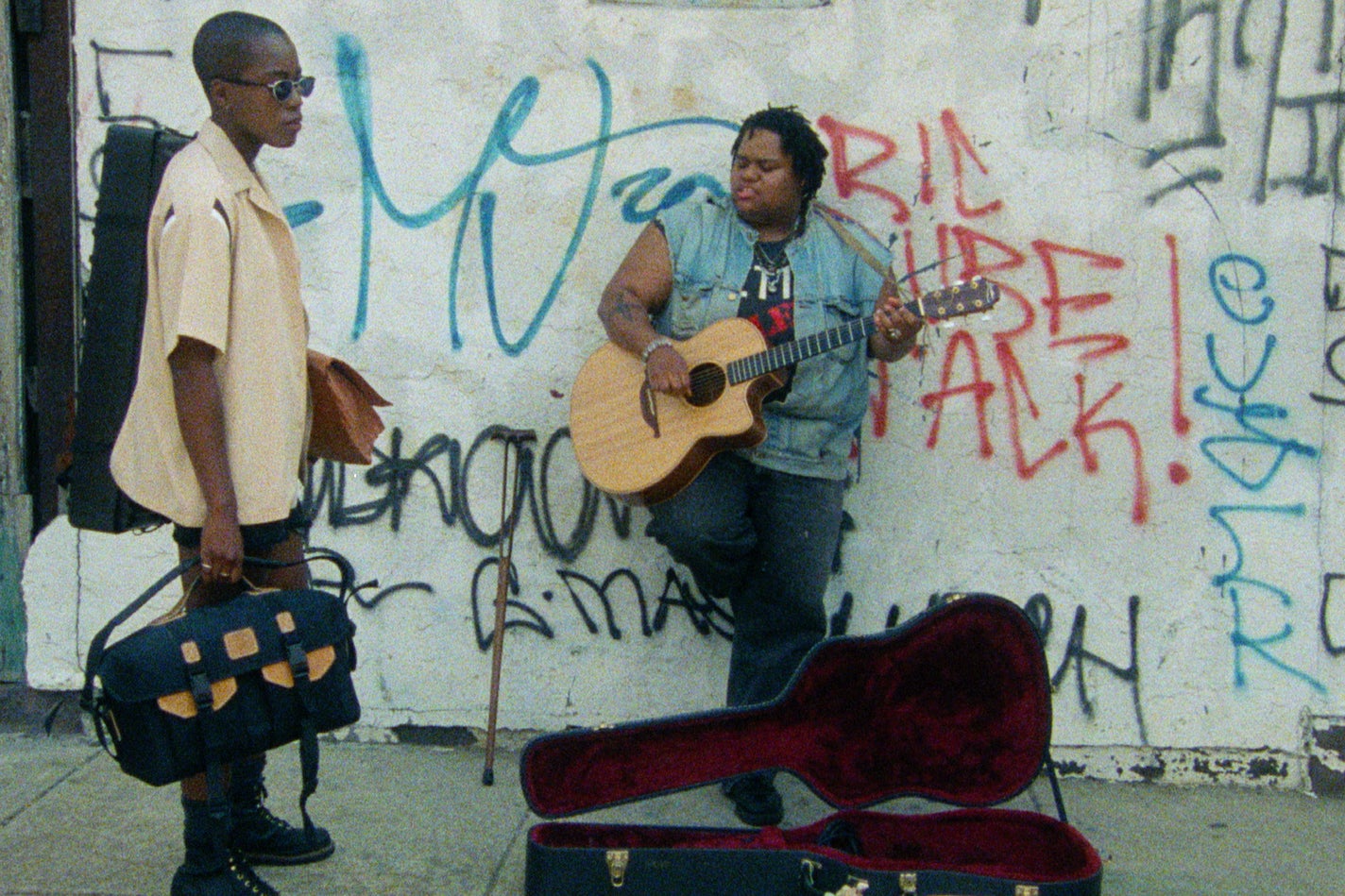 Two Black women standing on the street, one of them playing an acoustic guitar.