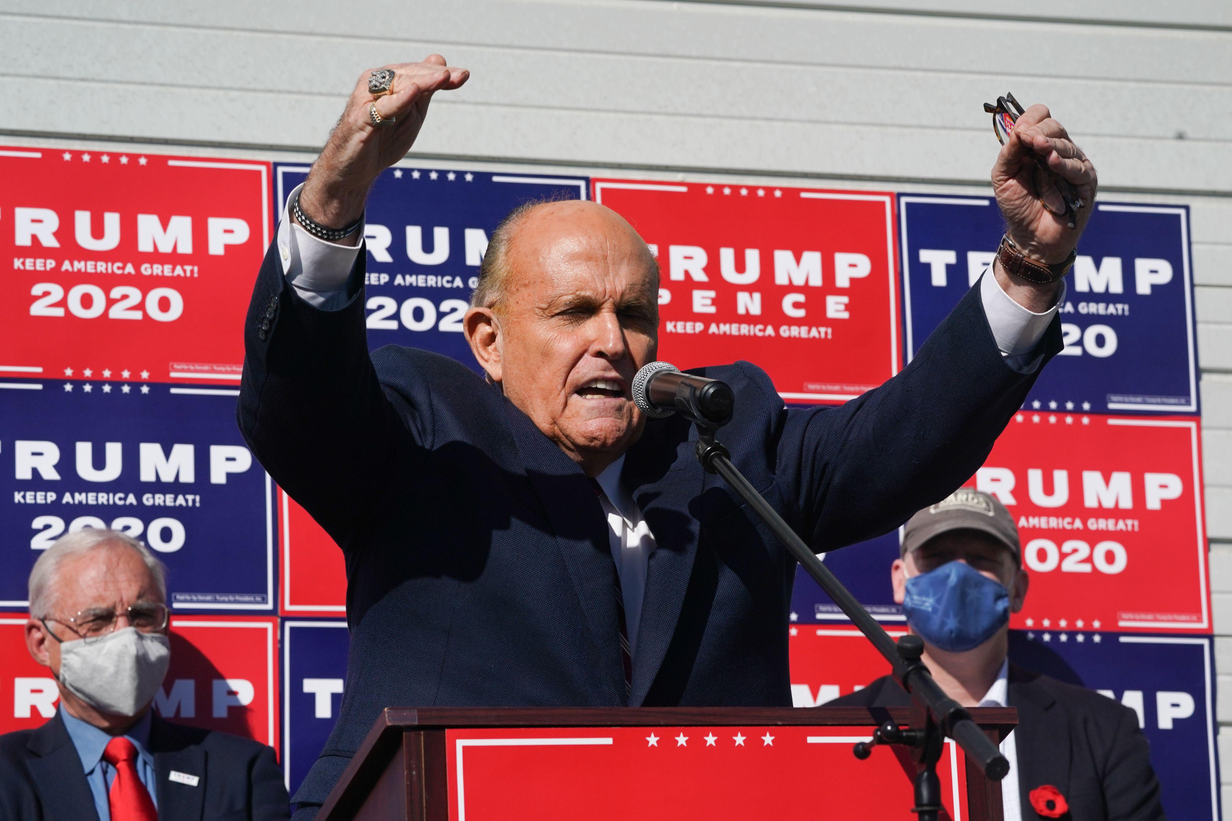 Giuliani stands at a podium in front of Trump 2020 signs and raises his hands, as two men stand in the background.