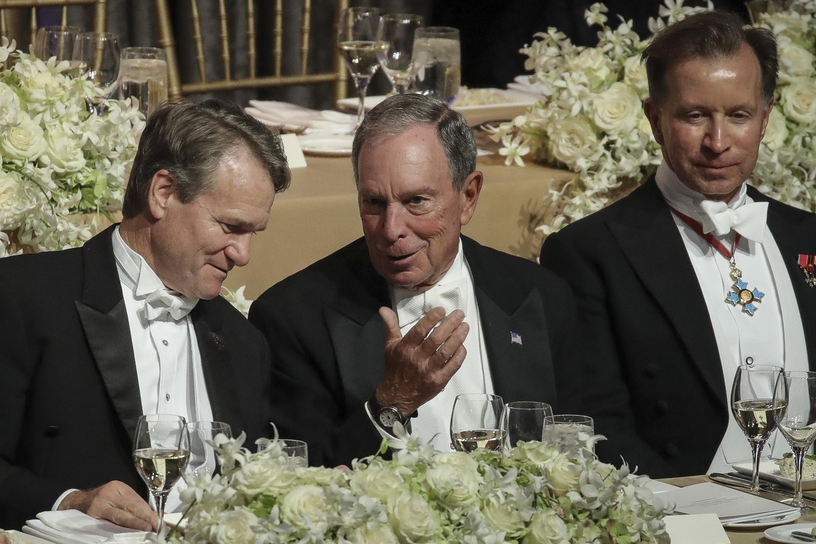 Michael Bloomberg, wearing white tie formal wear, talks at a banquet table