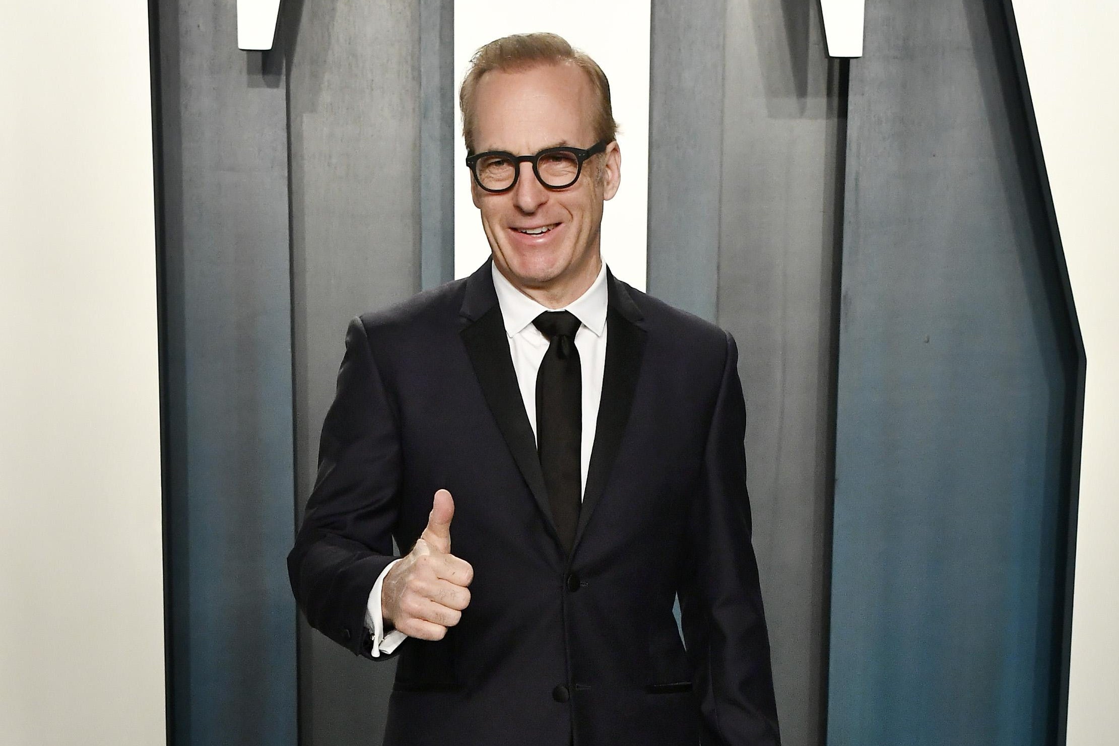 Bob Odenkirk in a black suit, standing in front of a Vanity Fair sign at their party, giving a thumbs up.