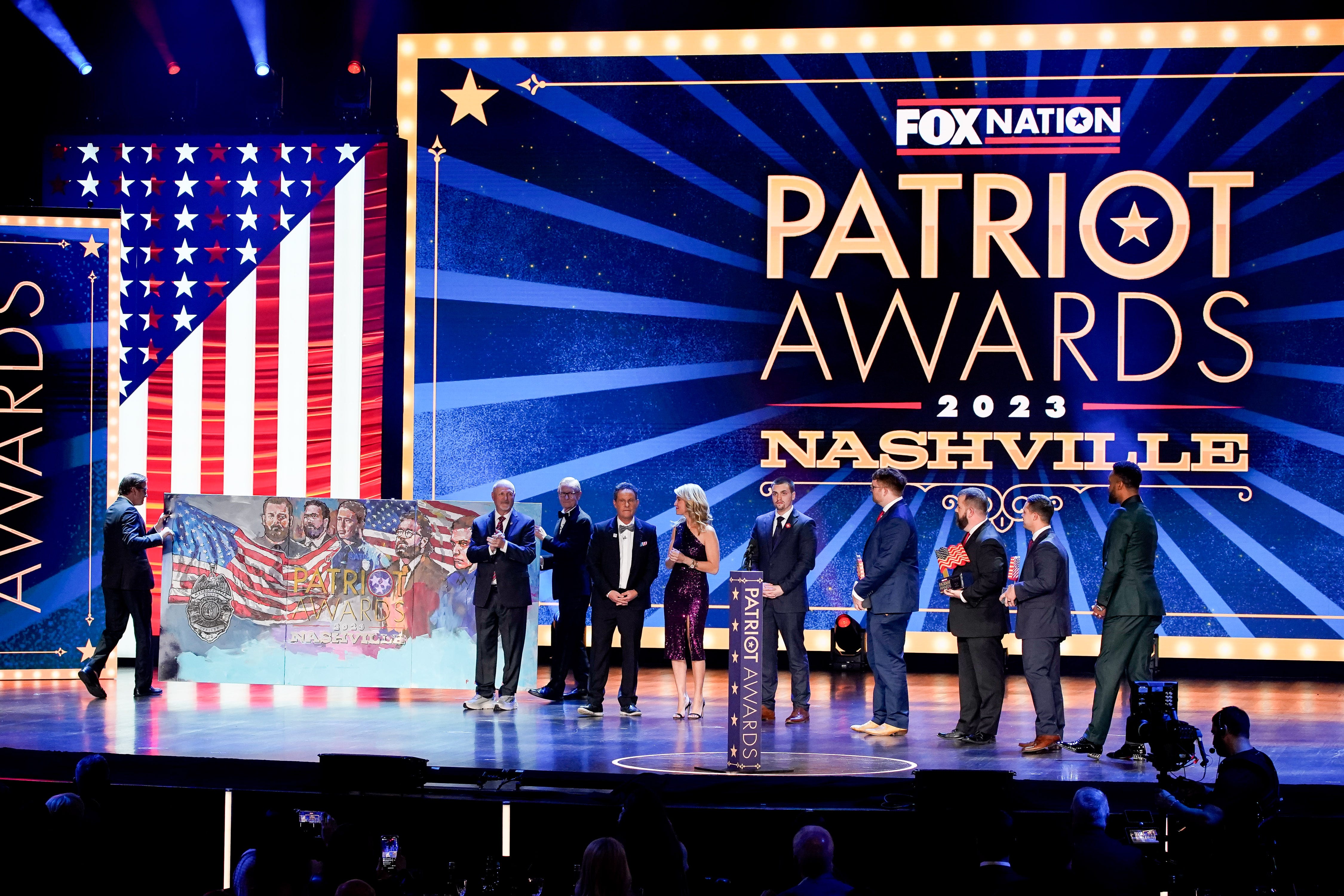 A brightly lit stage with the words "Patriot Awards 2023 Nashville" and an American flag behind a group of people receiving an award.