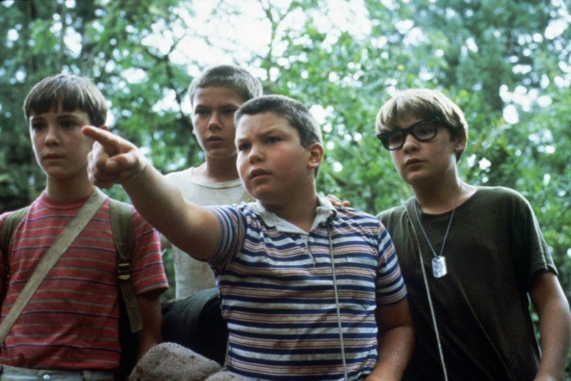 Four adolescent boys stand close together surrounded by trees looking past the camera. From left to right: Wil Wheaton, River Phoenix, Jerry O'Connell who is pointing at something, and Corey Feldman.