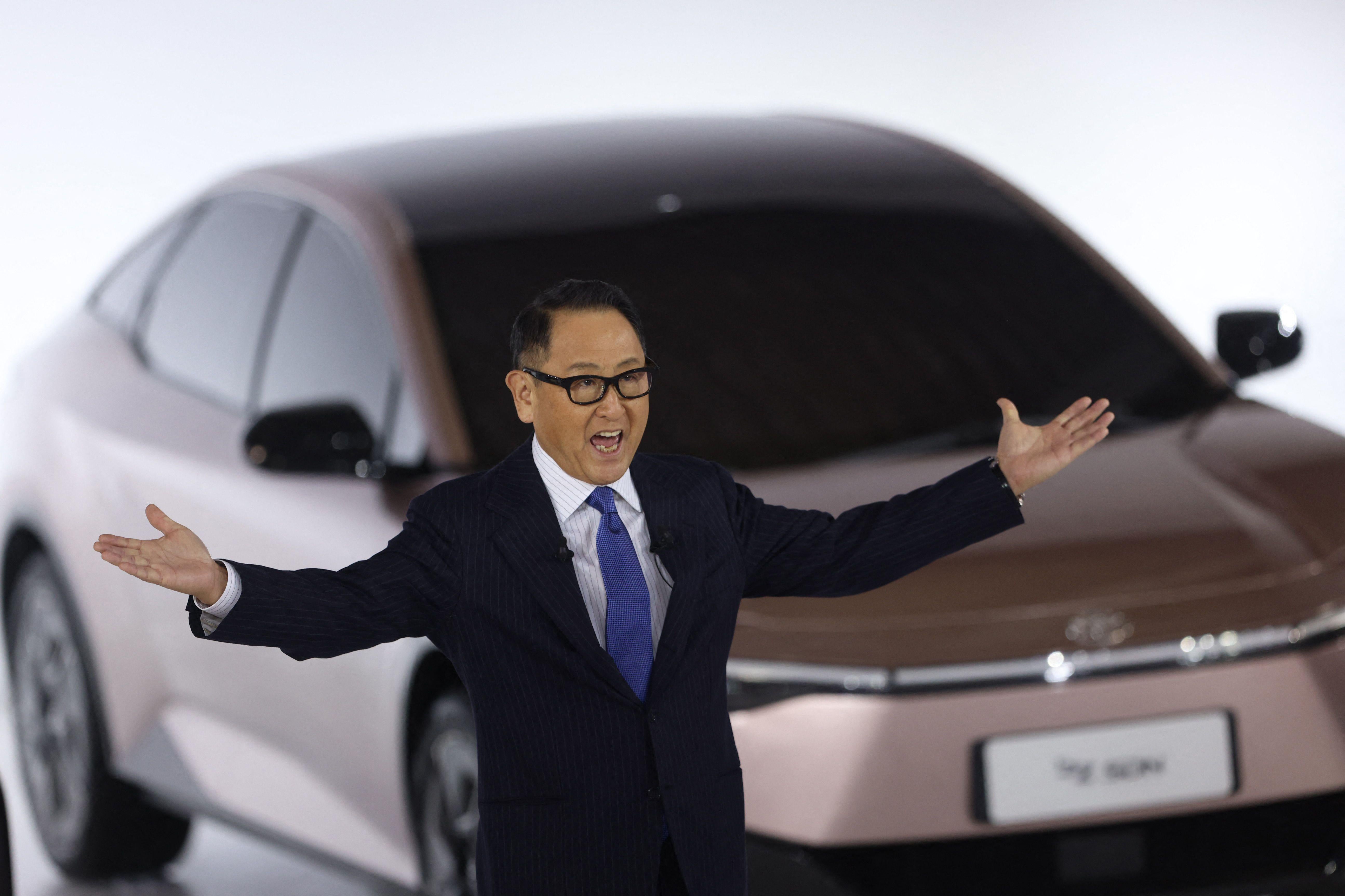 Akio Toyoda stretches out his hands while standing in front of a Toyota electric vehicle prototype.