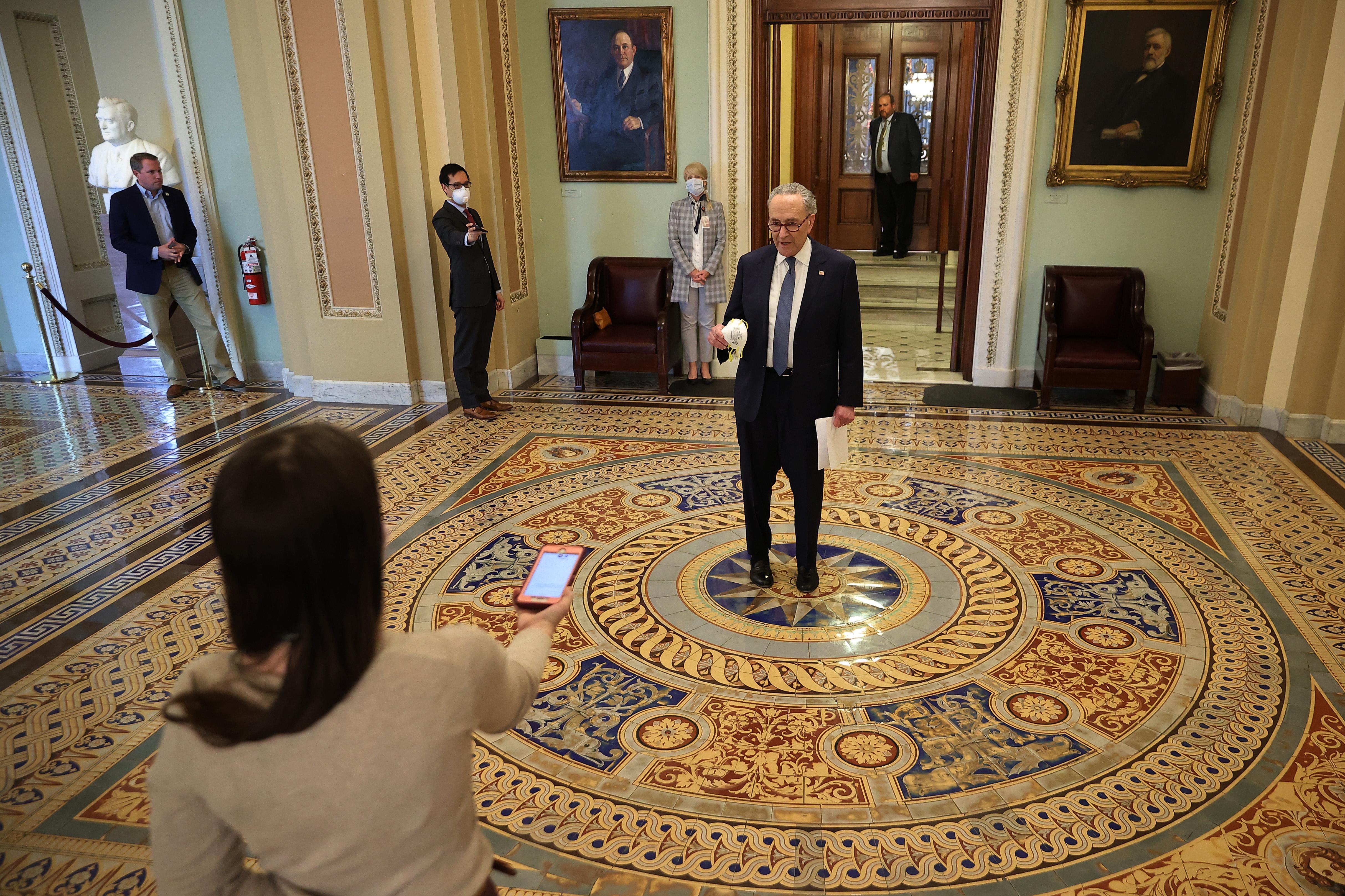 Schumer stands in the center of a room speaking to a reporter several feet away, recording the interview on a phone