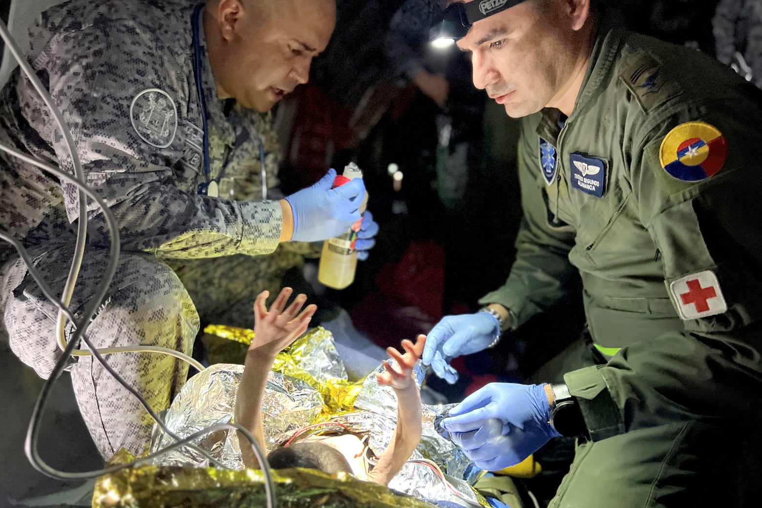 Two men in military uniforms, medics, tend to a baby whose arms are in the air. The baby is hooked up to various tubes and is in a makeshift hospital bed.