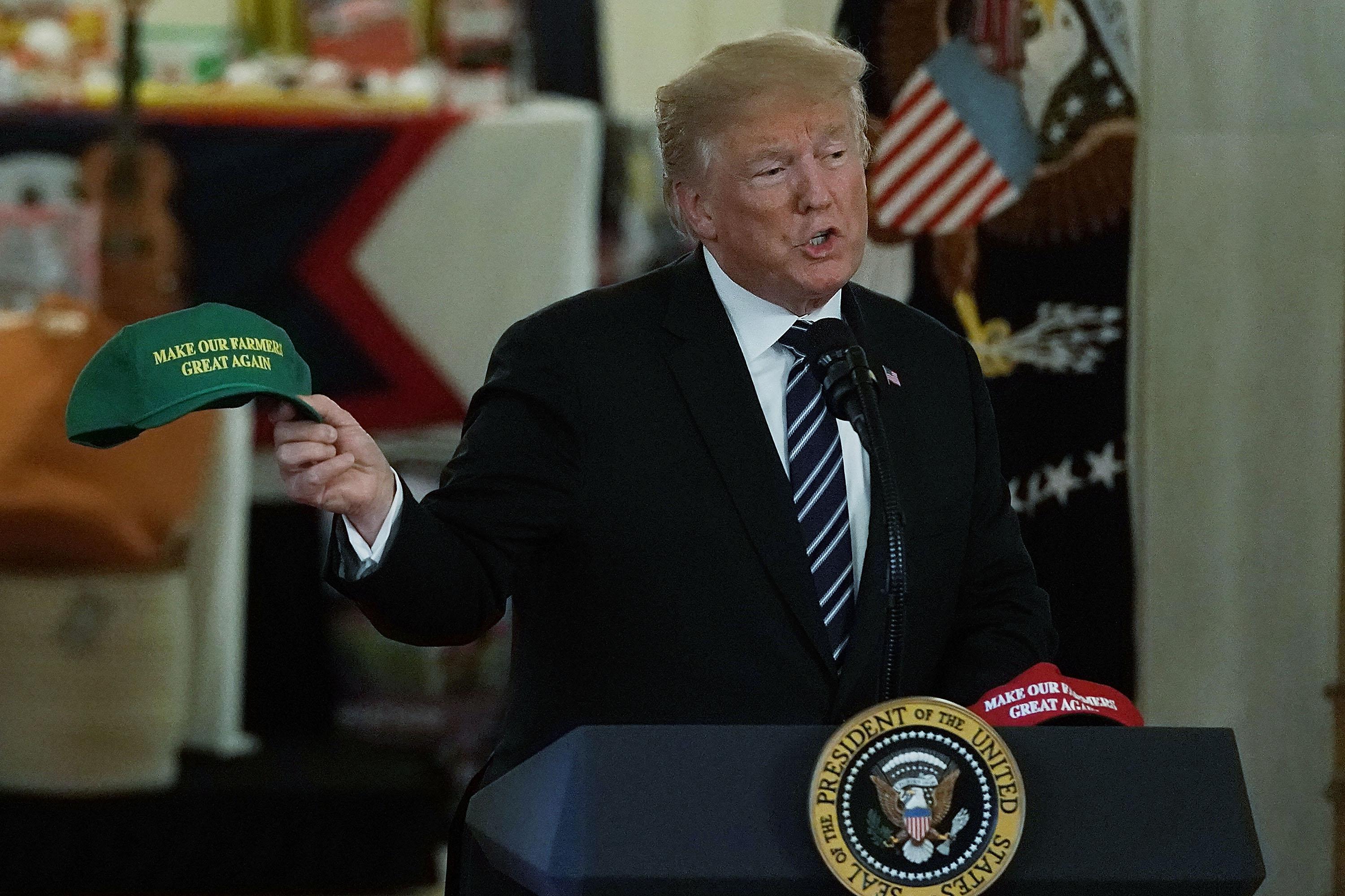 Donald Trump holds up a "Make Our Farmers Great Again" hat as he speaks at a podium.