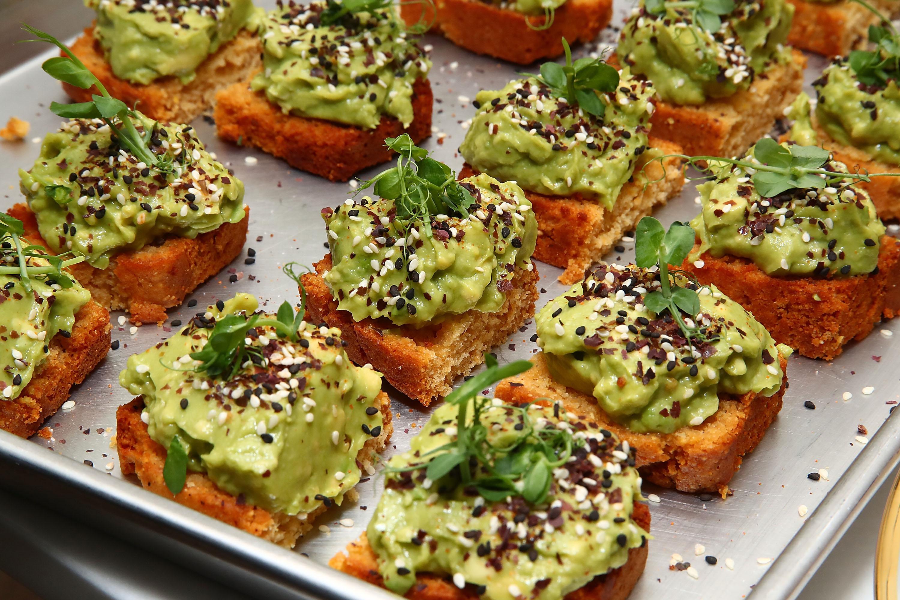 Rows of avocado toast sprinkled with sesame seeds sit on a metal sheet.