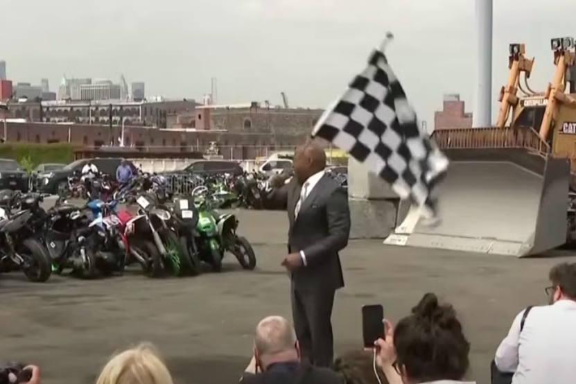 Eric Adams waves a checkered flag as a bulldozer approaches a line of dirt bikes.