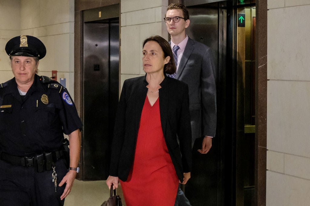 Hill, wearing a red desk and jacket, walks out of an elevator behind a police officer.