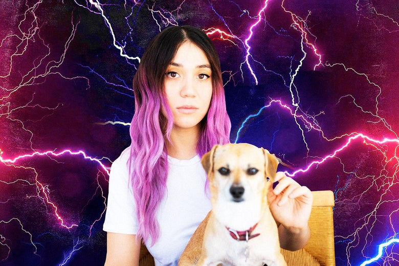 A woman with dyed purple hair wearing a white T-shirt faces the camera. She has a brown and white dog with a red collar on her lap, and she sits on a yellow chair. The background behind her is purple with lots of thin blue lightning bolts across it. 