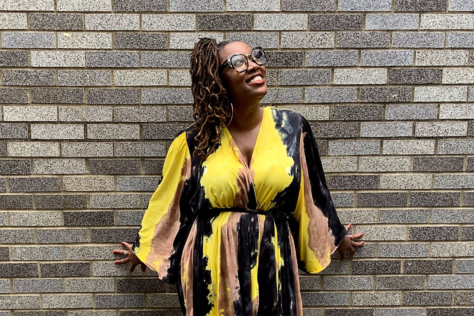Philyaw in a colorful dress smiling and looking skyward as she stands against a gray brick wall