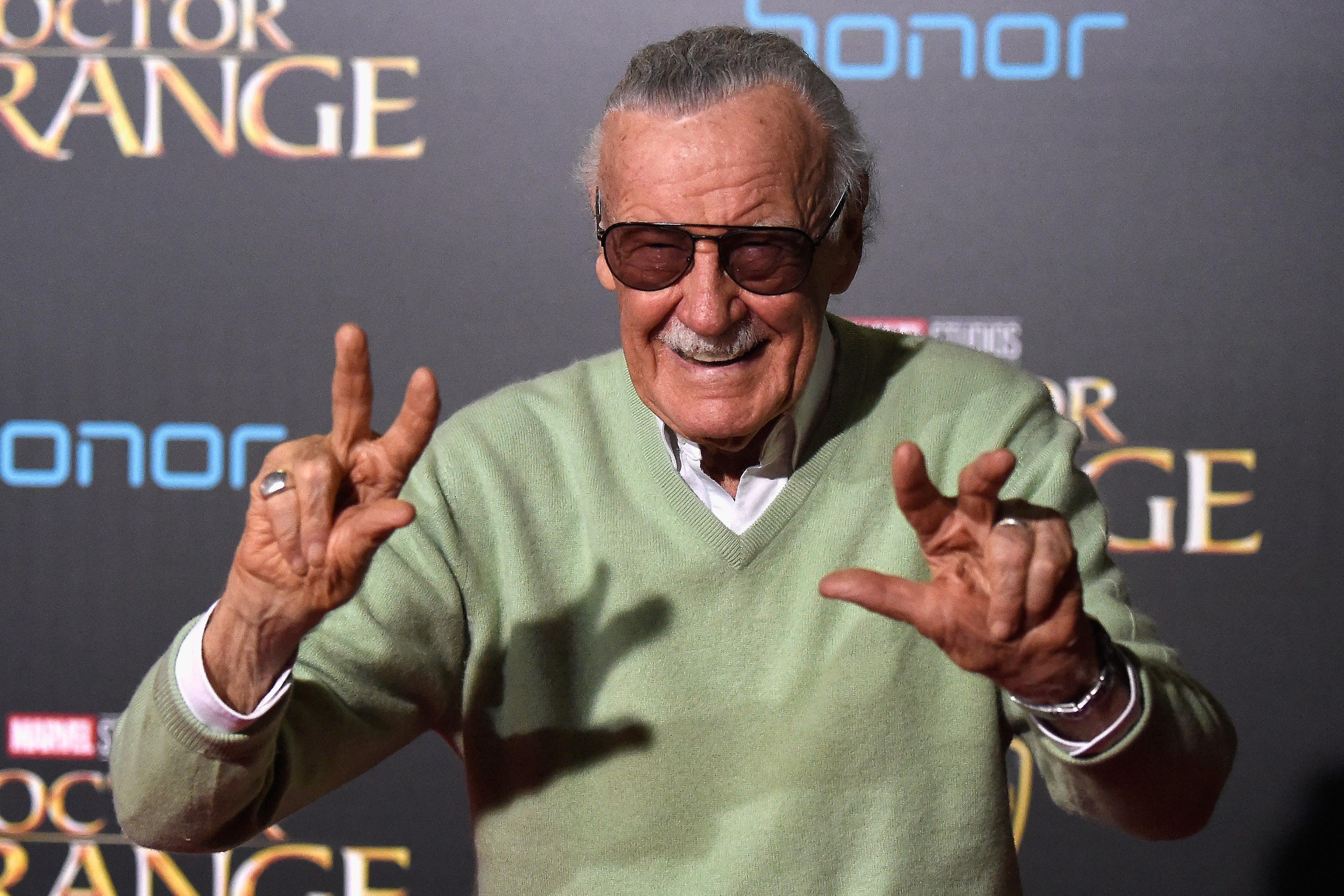 Stan Lee stands with two fingers raised on each hand.