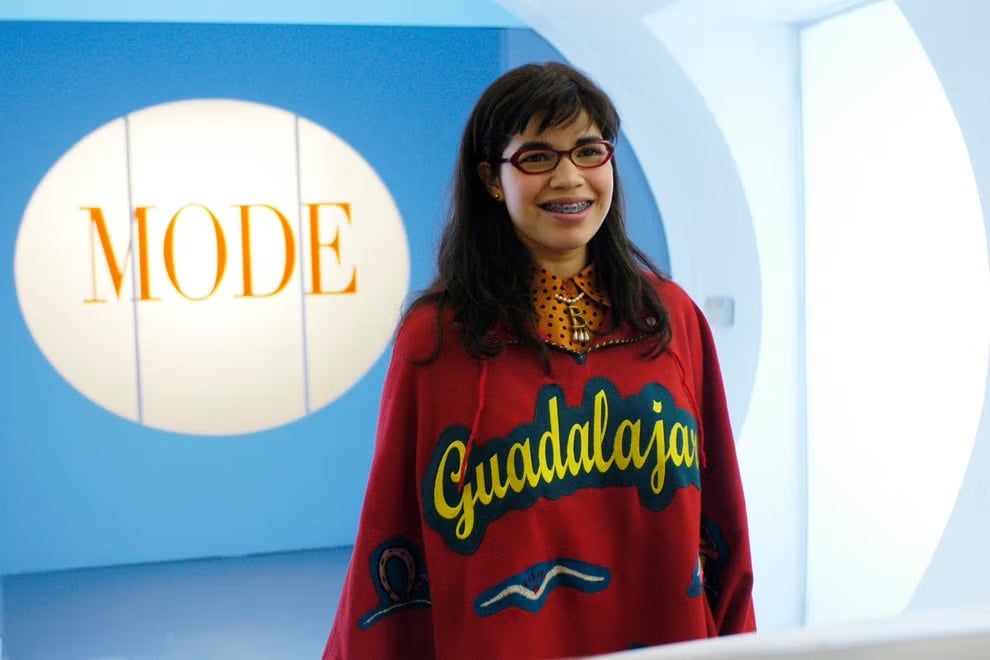 Young brunette with bangs, glasses, and braces, wearing a colorful poncho that says Guadalajara, stands in front of a check-in desk in a very chic, all-white waiting room.