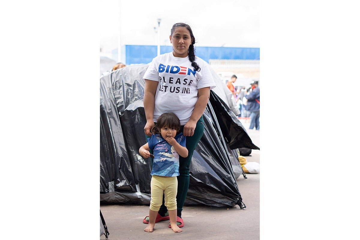 A woman in a T-shirt that says, "Biden, please let us in," stands behind a small child.