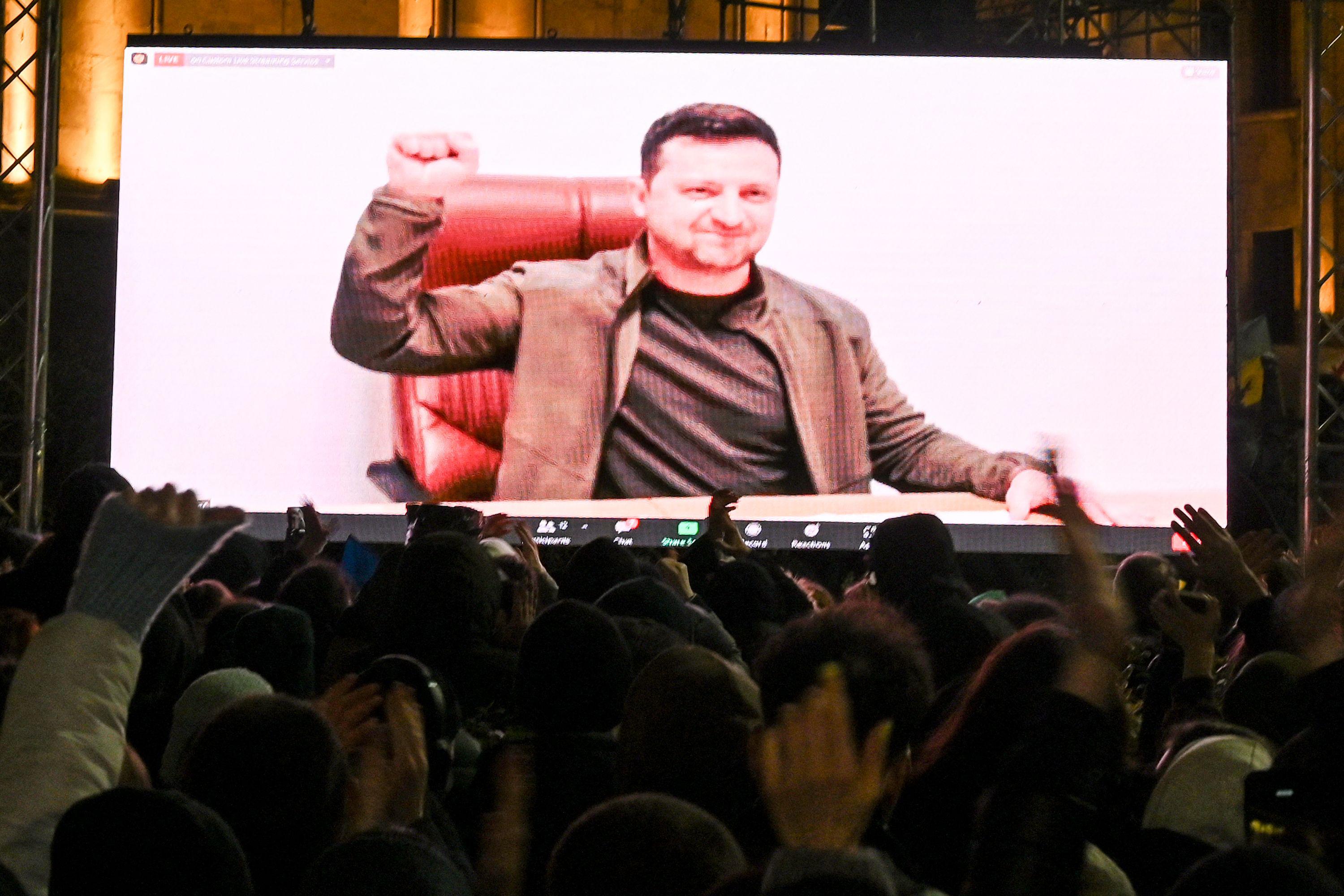 Zelensky raises a fist on a large TV screen in front of a crowd.