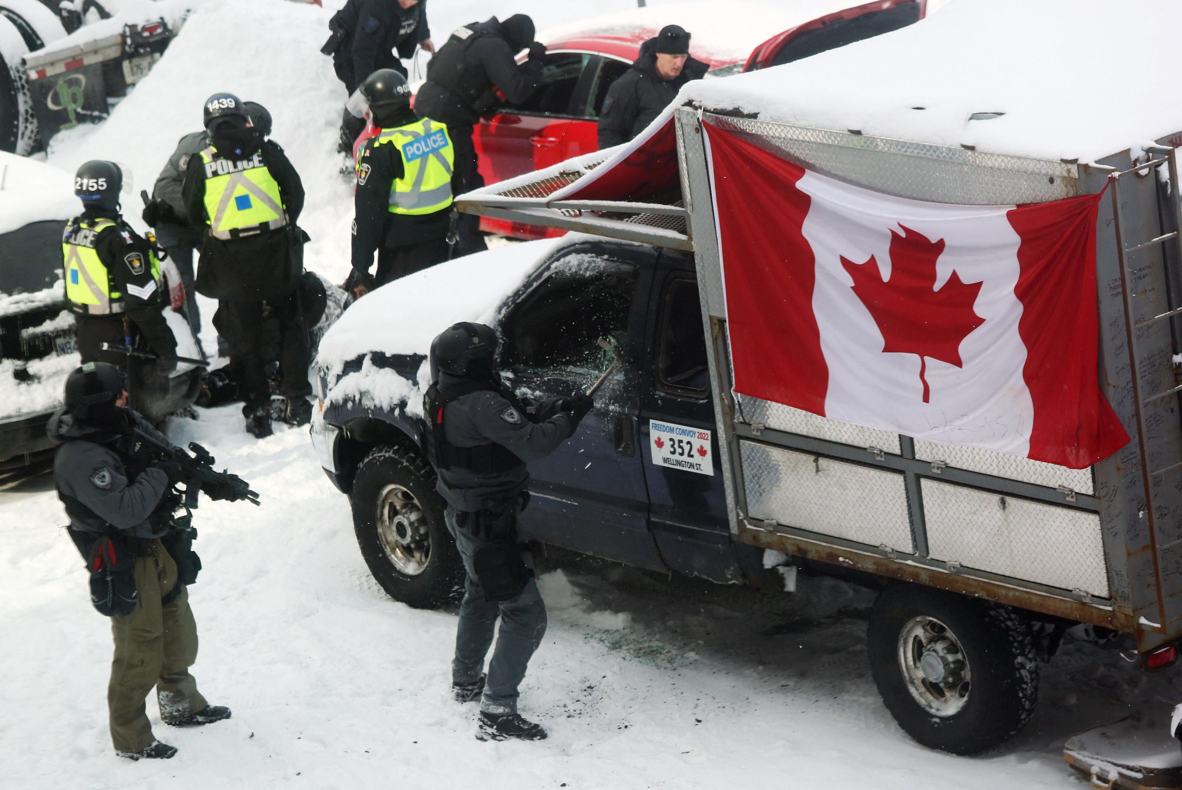 A small truck, draped with a Canadian flag, sits in the snow. It's surrounded by people in helmets and police vests. One person breaks the truck's driver-side window while someone stands behind them holding a rifle.