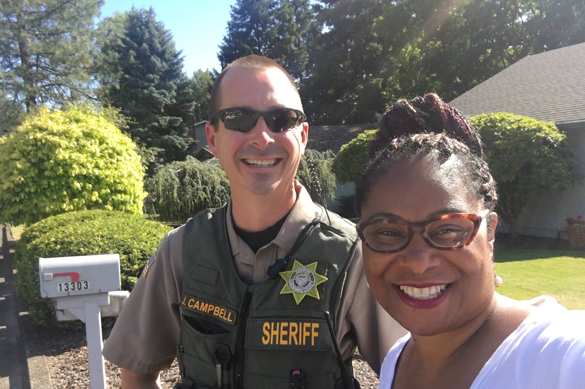 A uniformed police officer bearing the name tag J. Campbell, smiling next to Janelle Bynum in front of a house.