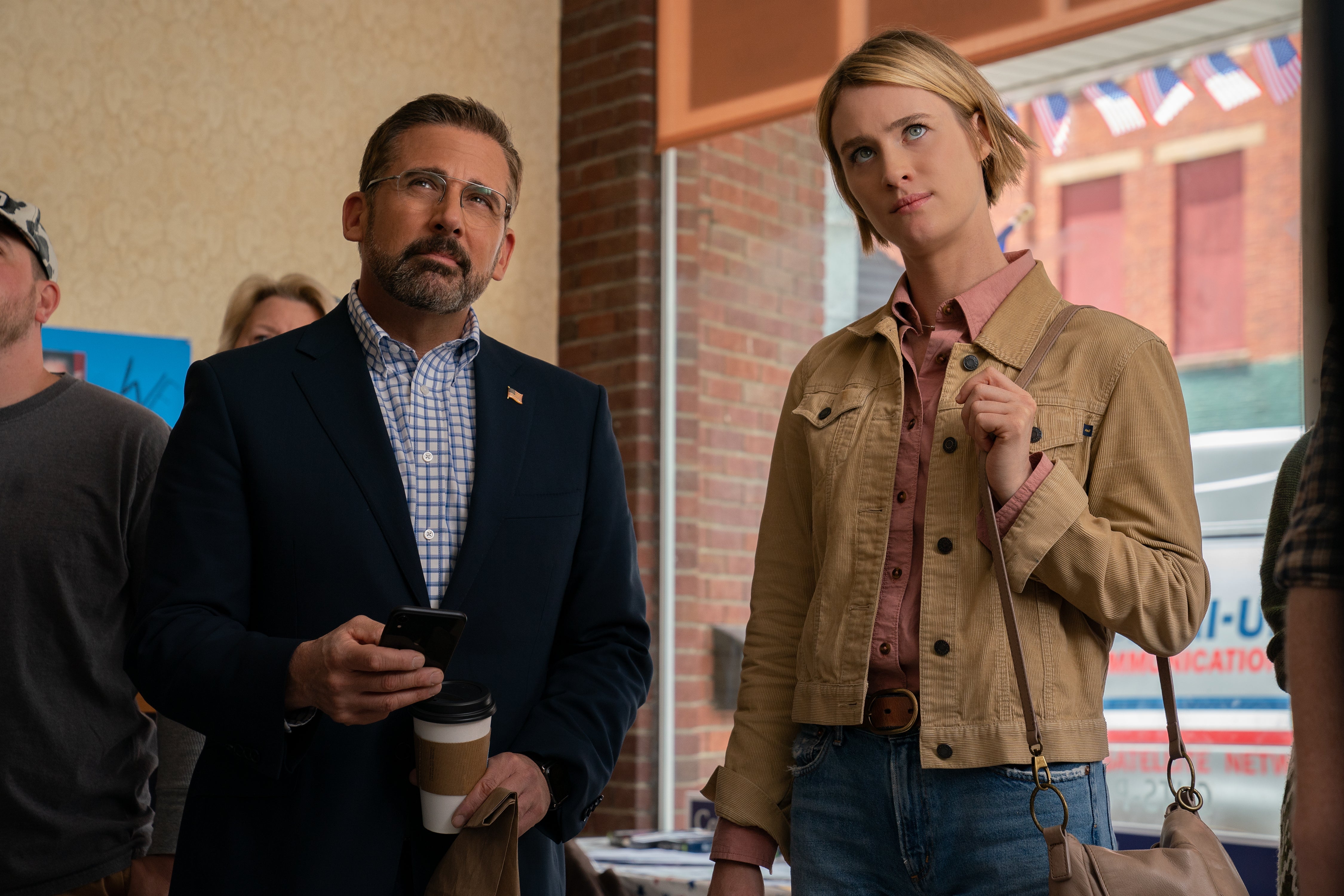 Carell wears a blazer with an American flag pin and holds a smartphone and a to-go cup of coffee. Mackenzie Davis looks comfortable in blue jeans. Neither seems to be happy with what they're watching.
