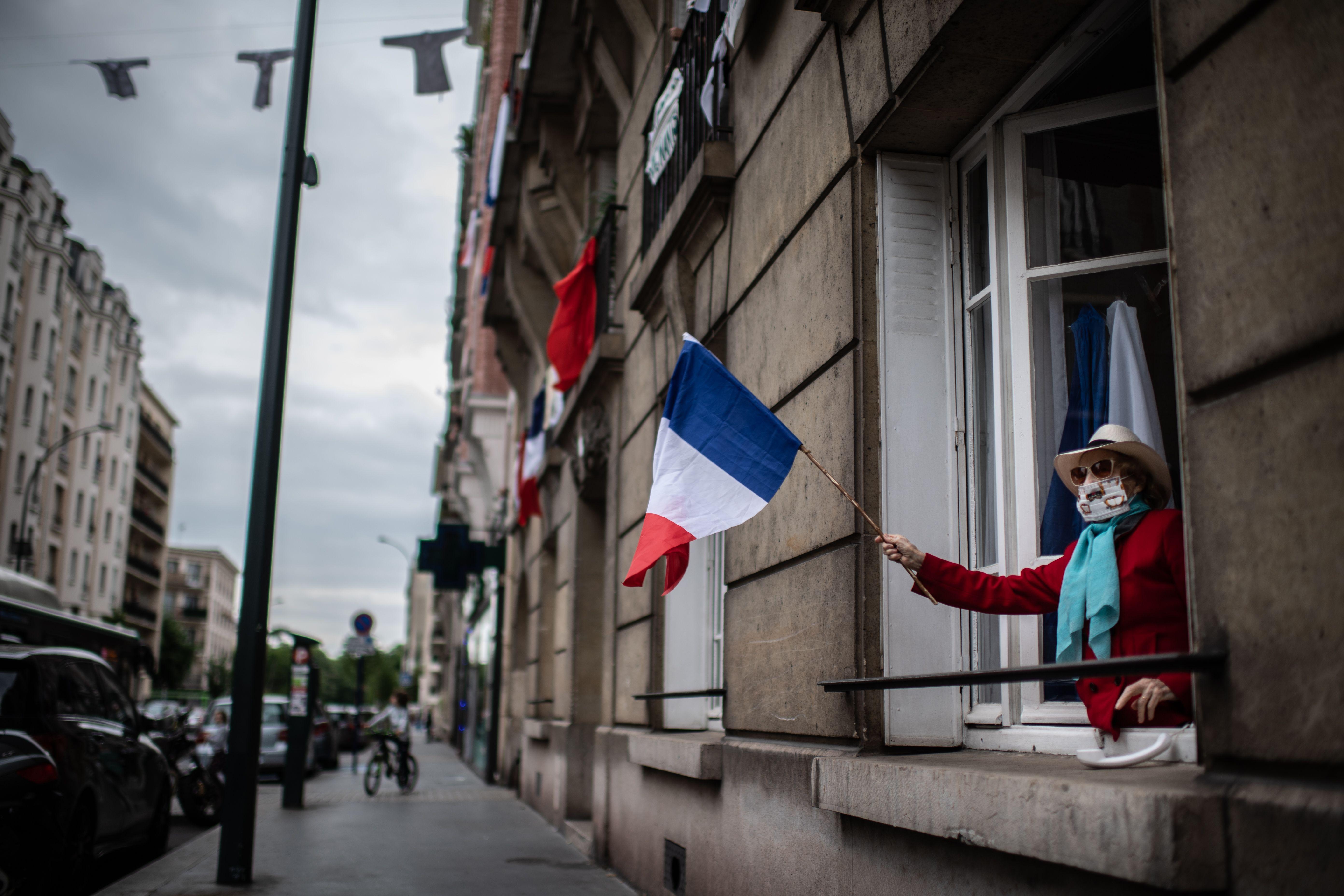 A woman wearing a mask leans out her first floor window and waves the French national flag to show support to healthcare employees.