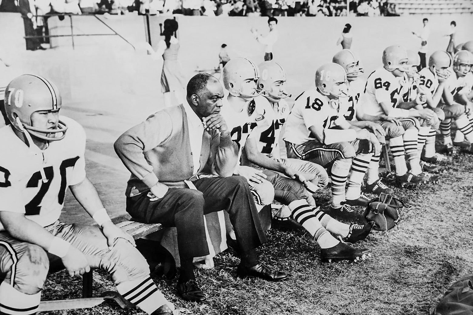 Washington as an older man in a cardigan and slacks sitting thoughtfully on a bench beside young uniformed football players during a game