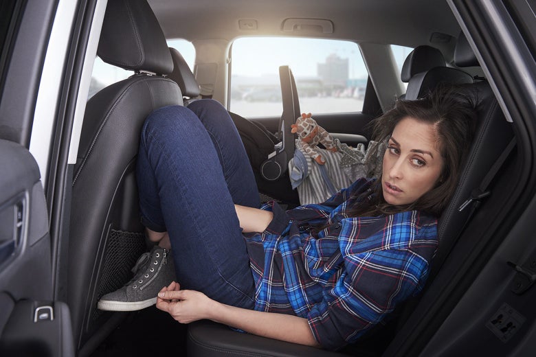 Catherine Reitman sits awkwardly in a car's rear, driver-side passenger seat in this still from Workin' Moms.
