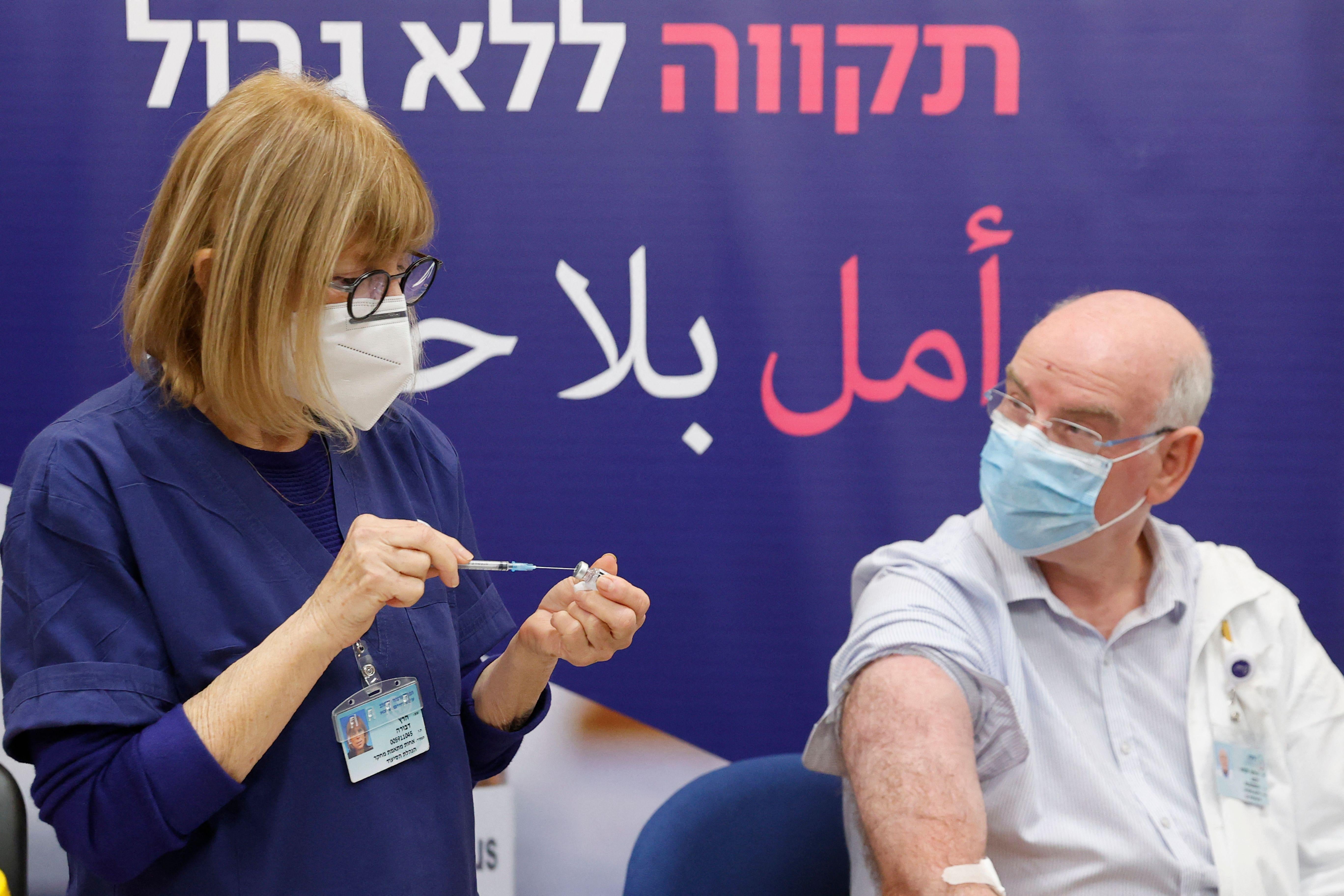 A nurse holds a syringe to a vial next to a man in a mask sitting with his right arm exposed