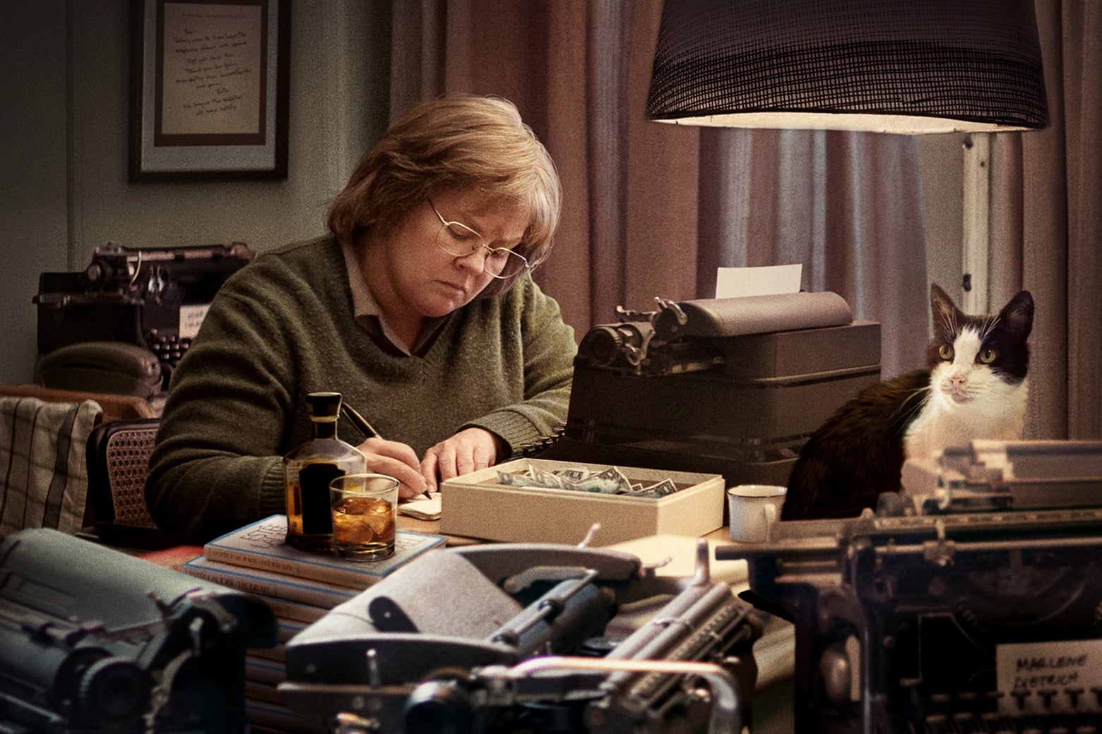 In a scene from Can You Ever Forgive Me? Lee Israel, played by Melissa McCarthy, sits at a desk forging a letter. There are several typewriters, a lamp, and a cat on the desk.