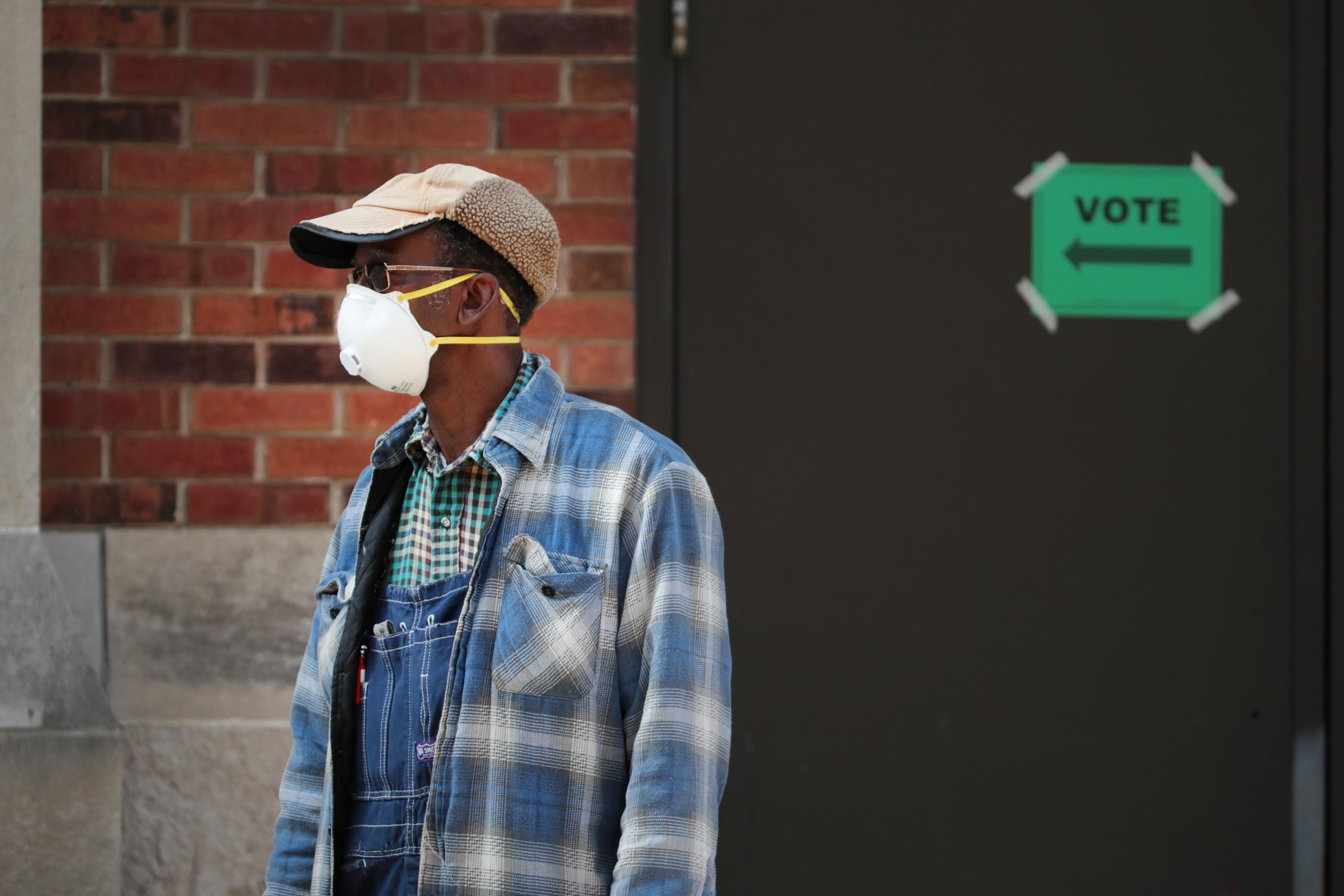 A person wears a face mask and stands near a door with a sign that says "Vote" and points an arrow to the left.