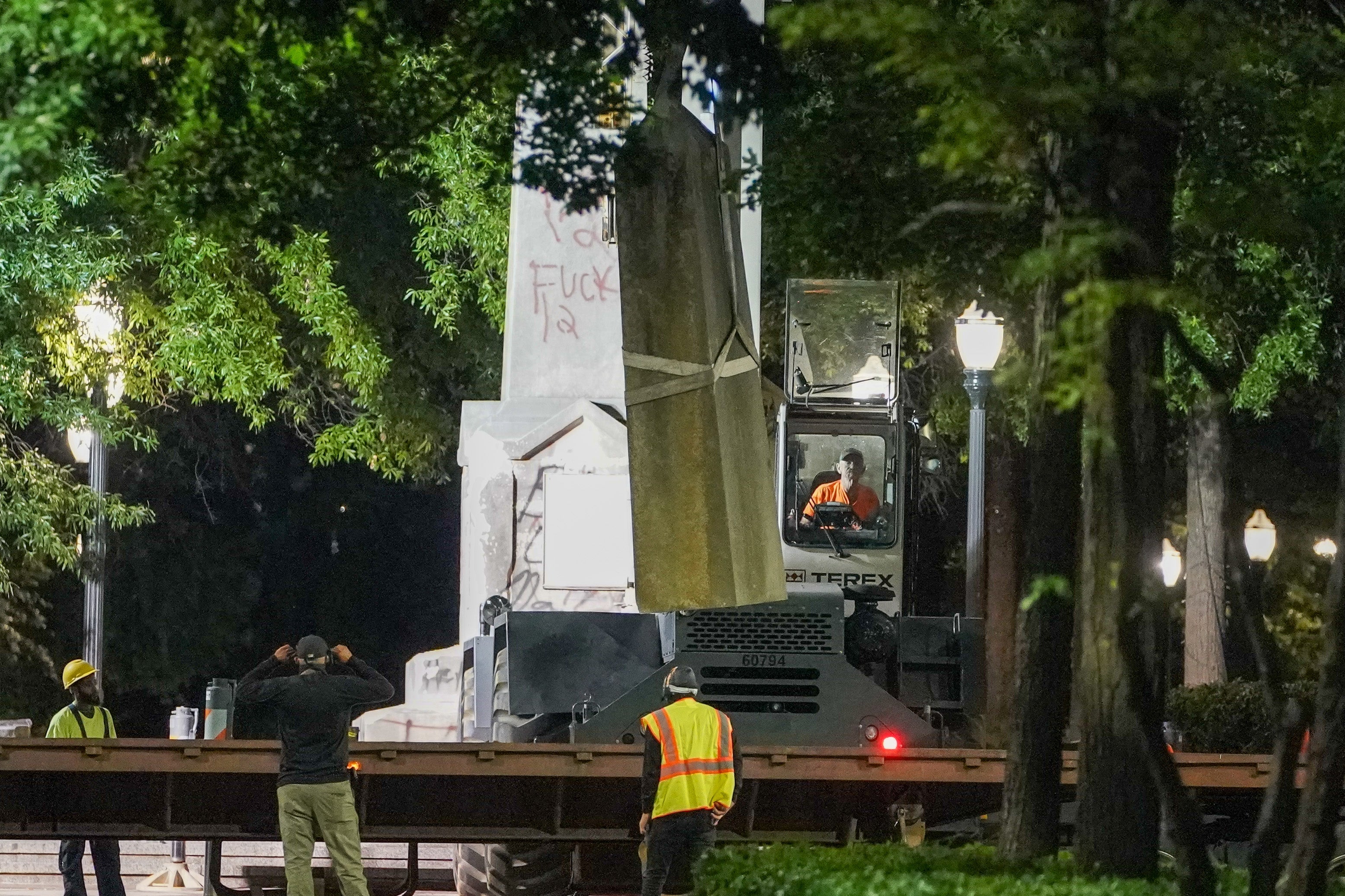 Construction workers with large machinery examine a graffiti-covered white obelisk.