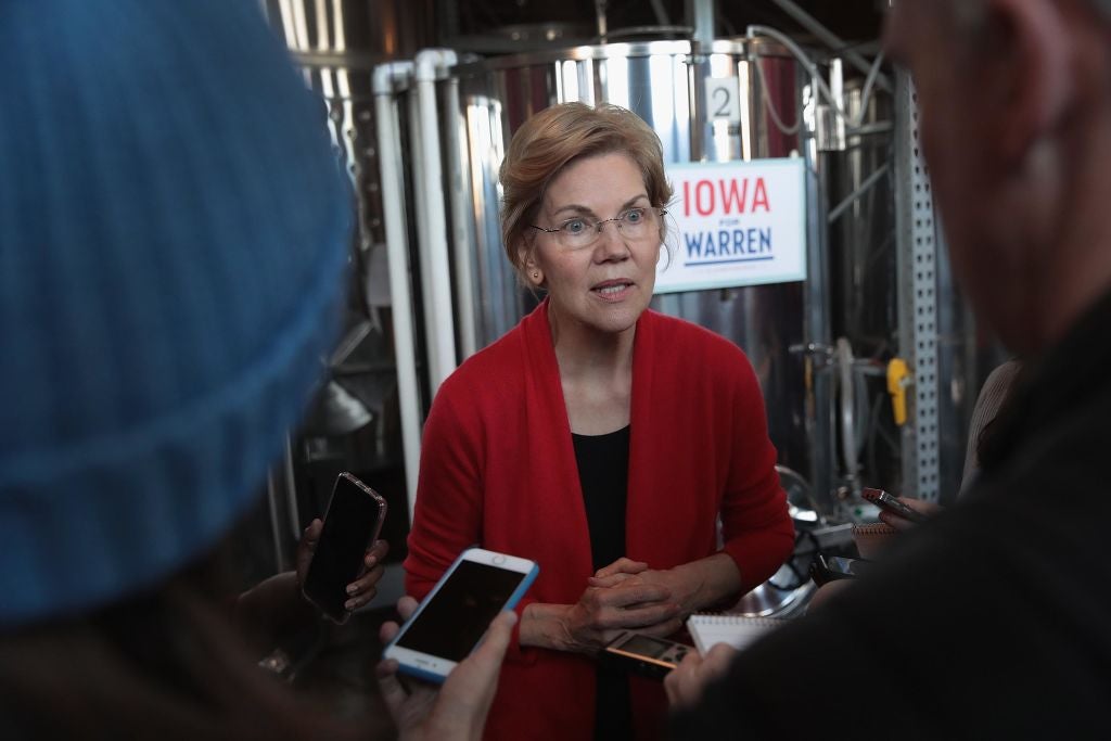 Warren, wearing a red blazer, speaks to two reporters while standing in front of a big old vat of wine.
