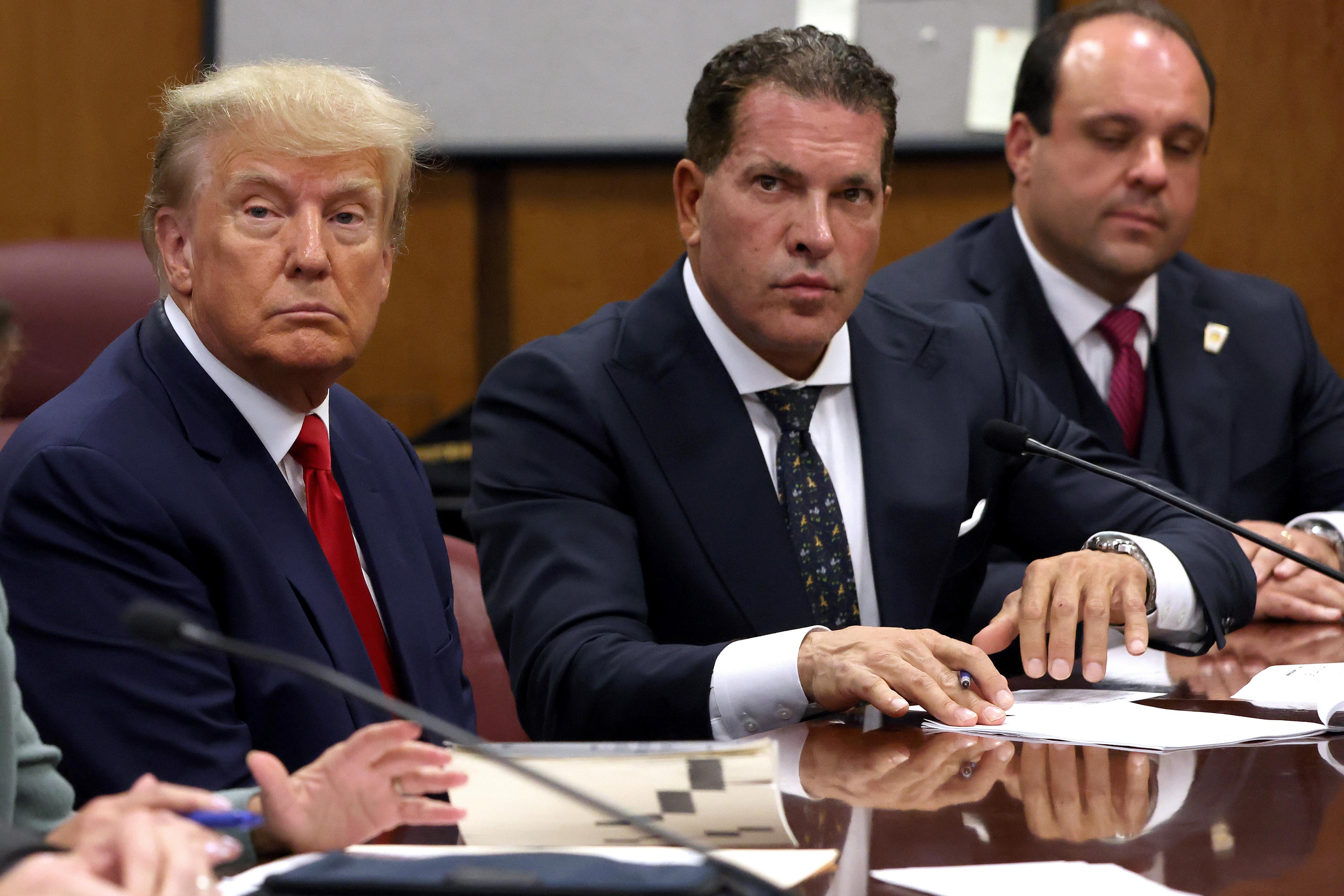 Trump, wearing a red tie, frowns and stares ahead while seated between his lawyers in court.