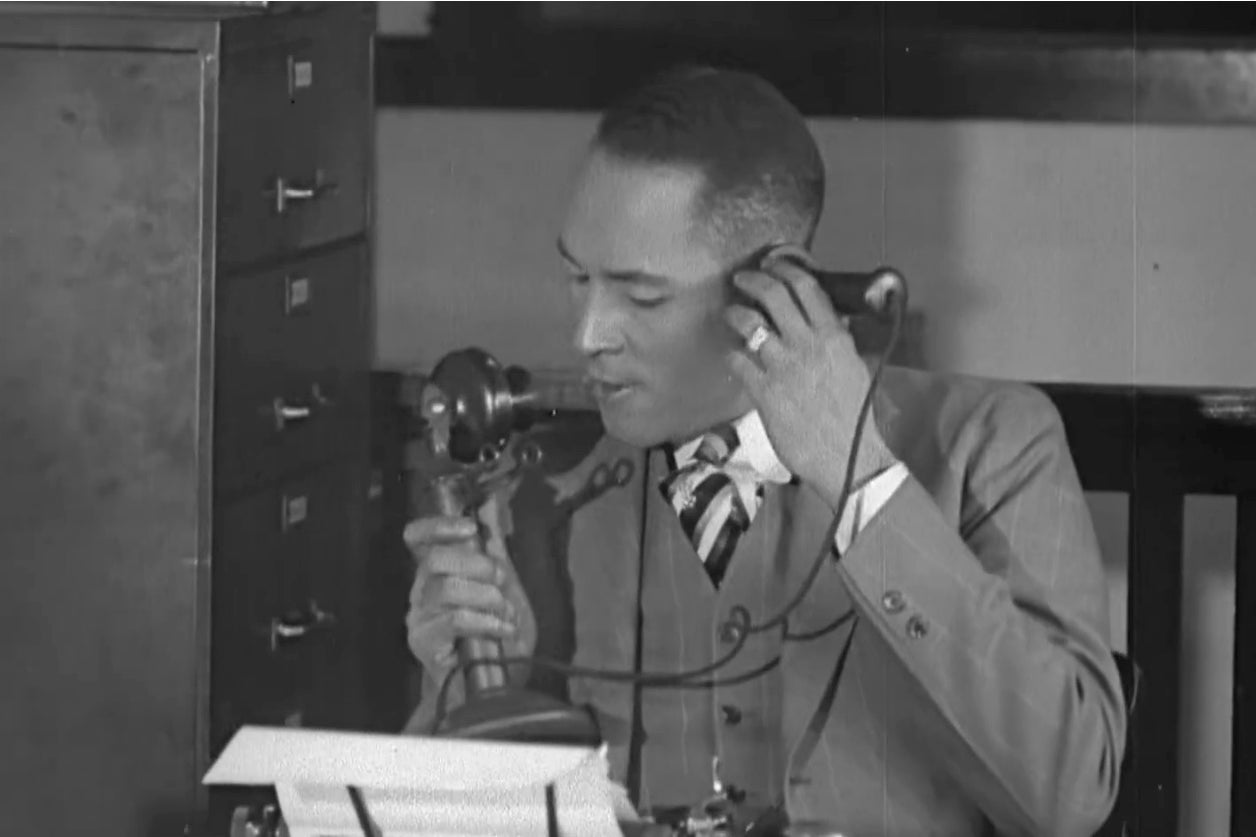 A Black man in a suit speaks on an old-timey telephone.