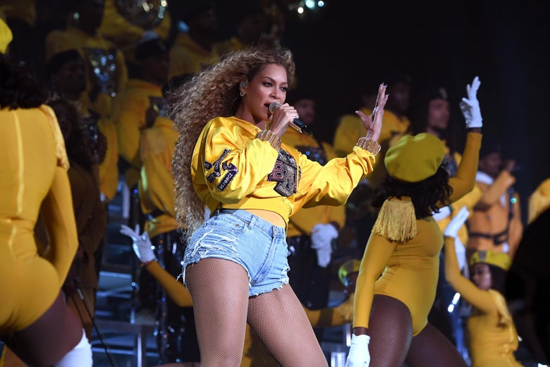 Beyoncé, dressed in a yellow and black shirt, performs on risers surrounded by dancers. 