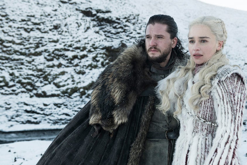 Kit Harington and Emilia Clarke, dressed in fur, stand in front of a snowy mountain landscape.