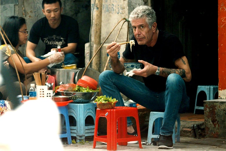 Anthony Bourdain sitting on a small stool eating noodles with chopsticks.