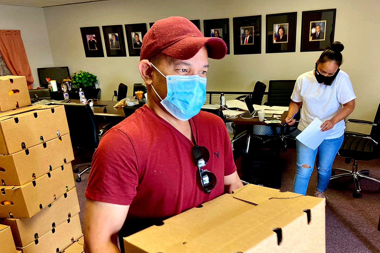 A man in a mask, red T-shirt, and red ball cap carries a cardboard box.