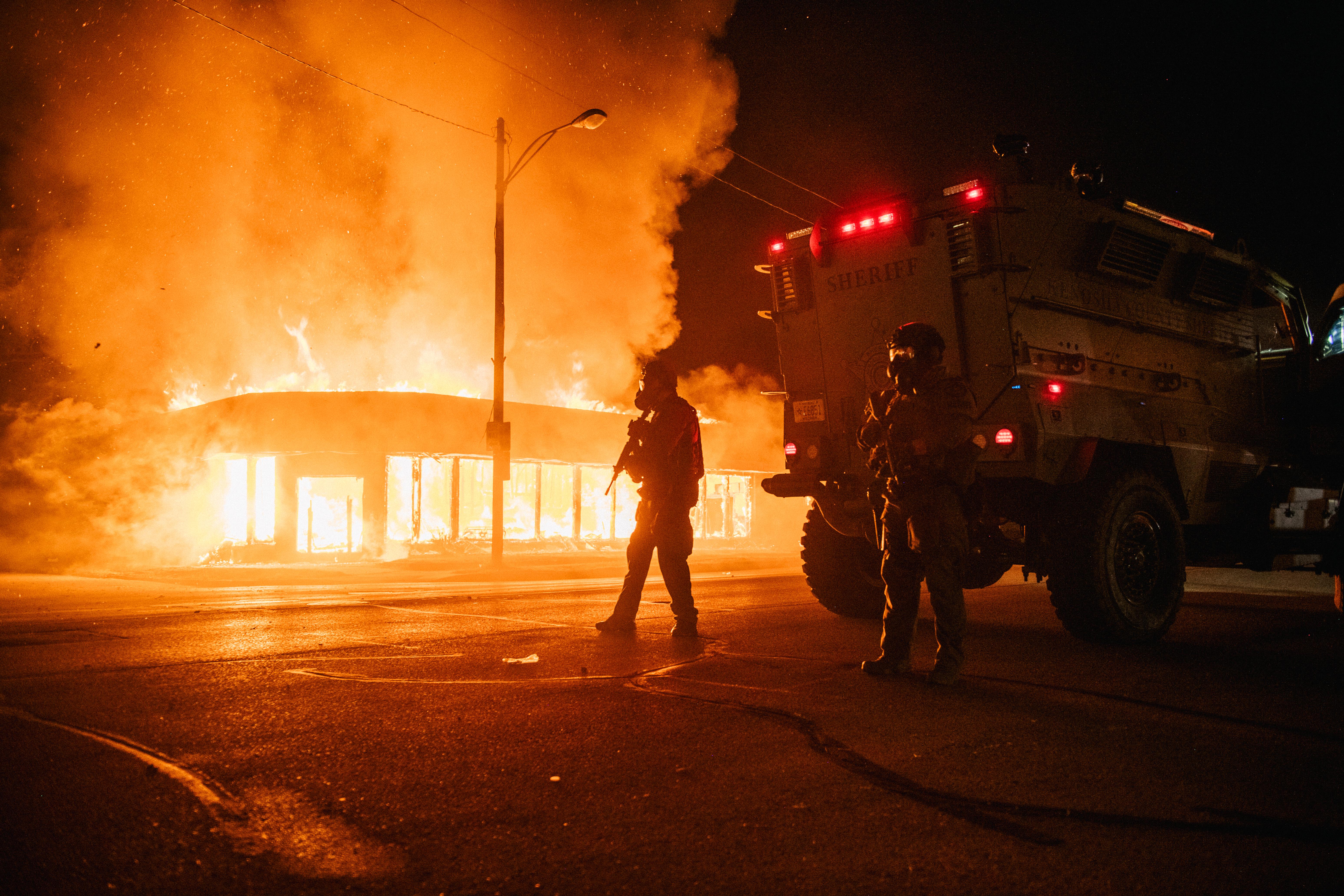 A police armored vehicle patrols an intersection with a building on fire in the background. 