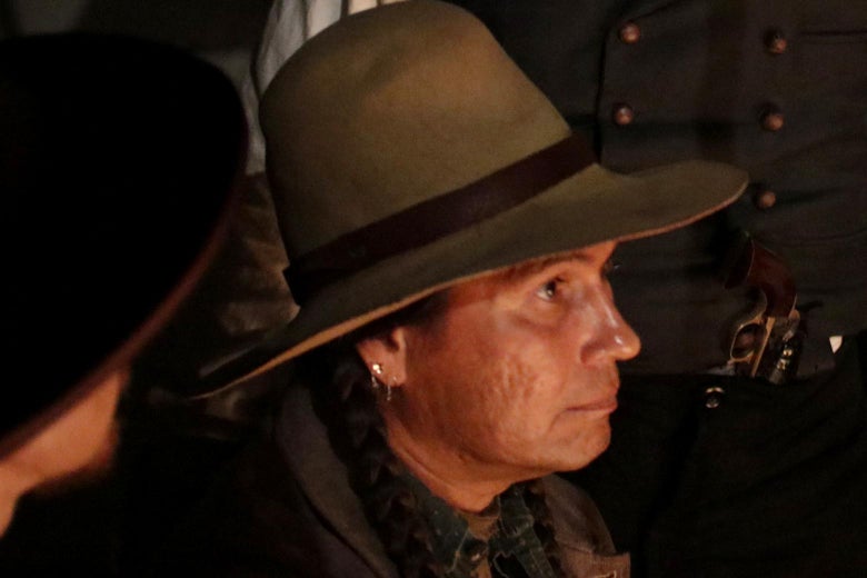 Mo Brings Plenty portraying Ottawa Jones wearing a hat and sitting by a fire.