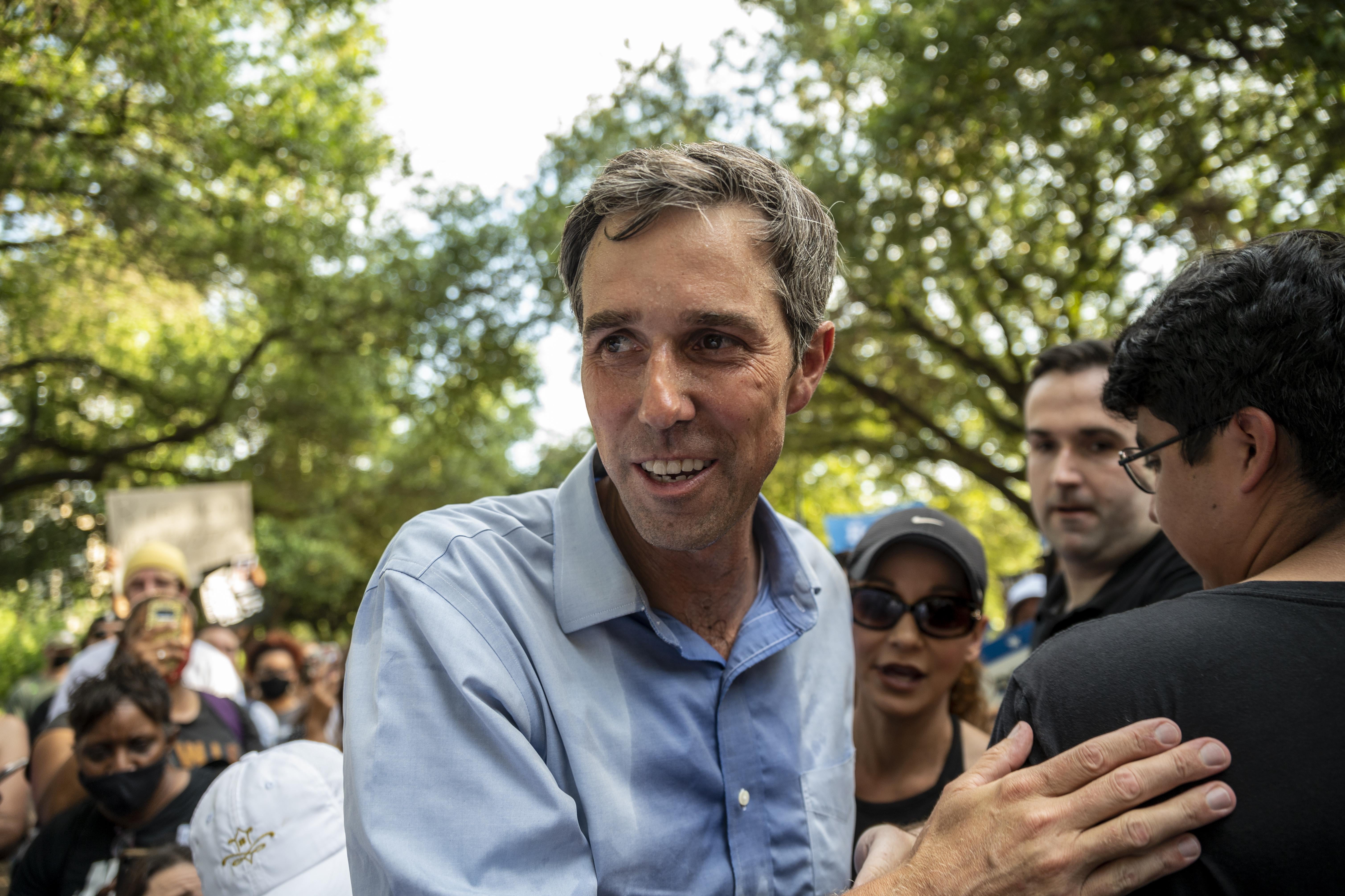 Beto in a button-down drenched in sweat smiles and pats someone's back as he walks through a crowd in a tree-lined area