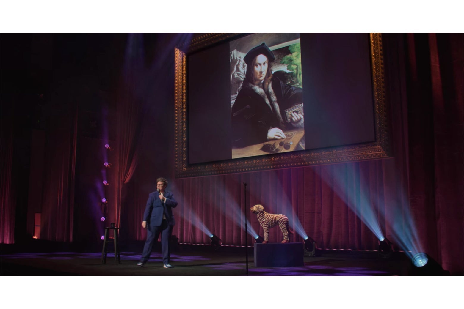 Hannah Gadsby onstage in front of a portrait of an angry-looking man dressed all in black.
