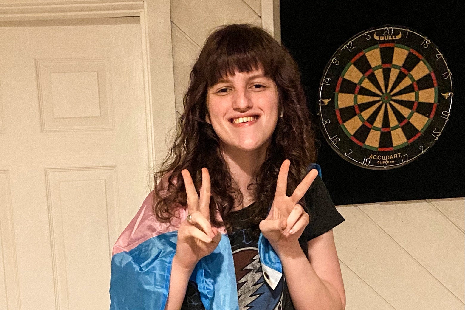 Esmée Silverman, smiling, holds her hands in peace signs and wears a transgender flag draped over her shoulders.