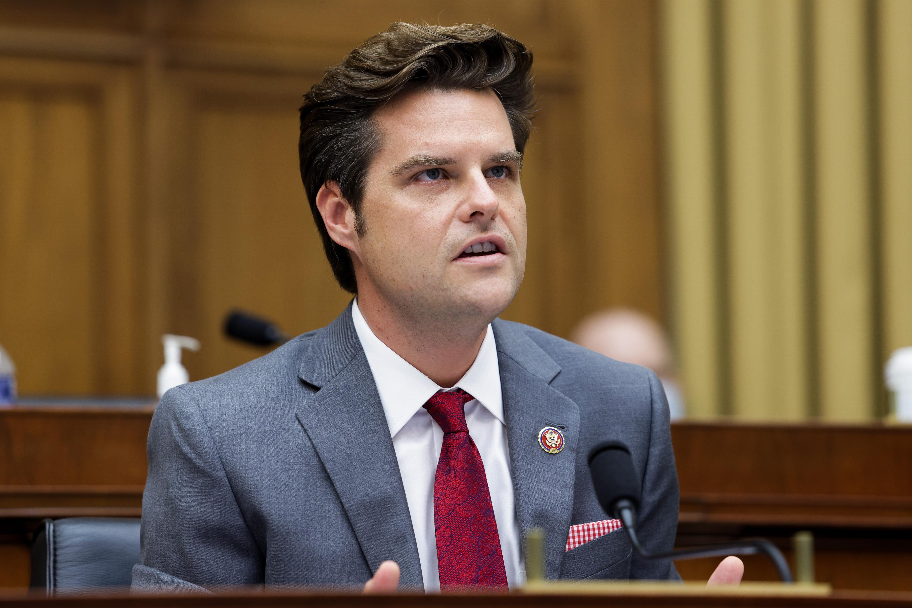 A man in a suit, tie, and congressional pin speaks in front of a microphone.