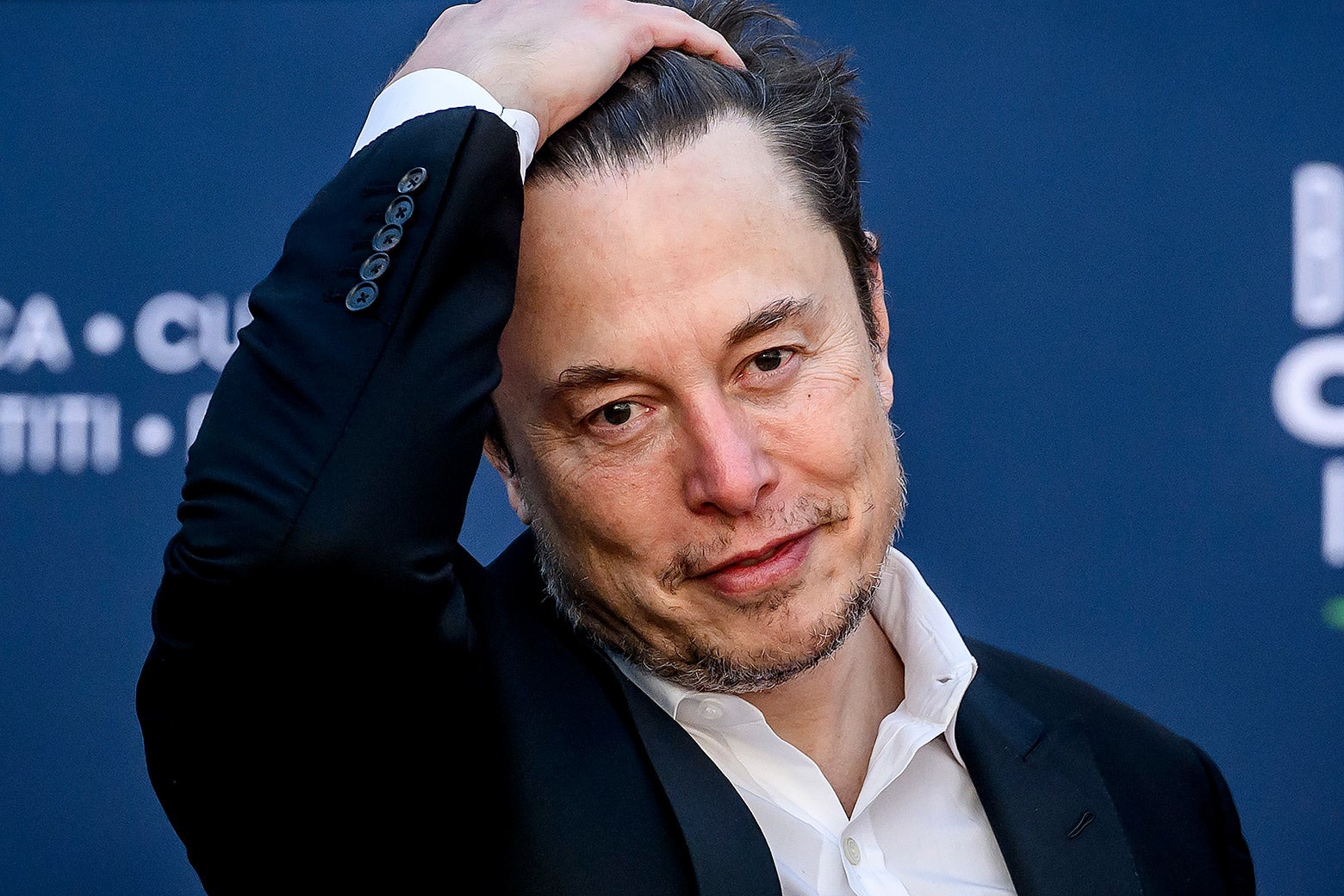 Musk, wearing a white shirt and navy blazer, runs his hand through his hair and looks mildly frustrated.