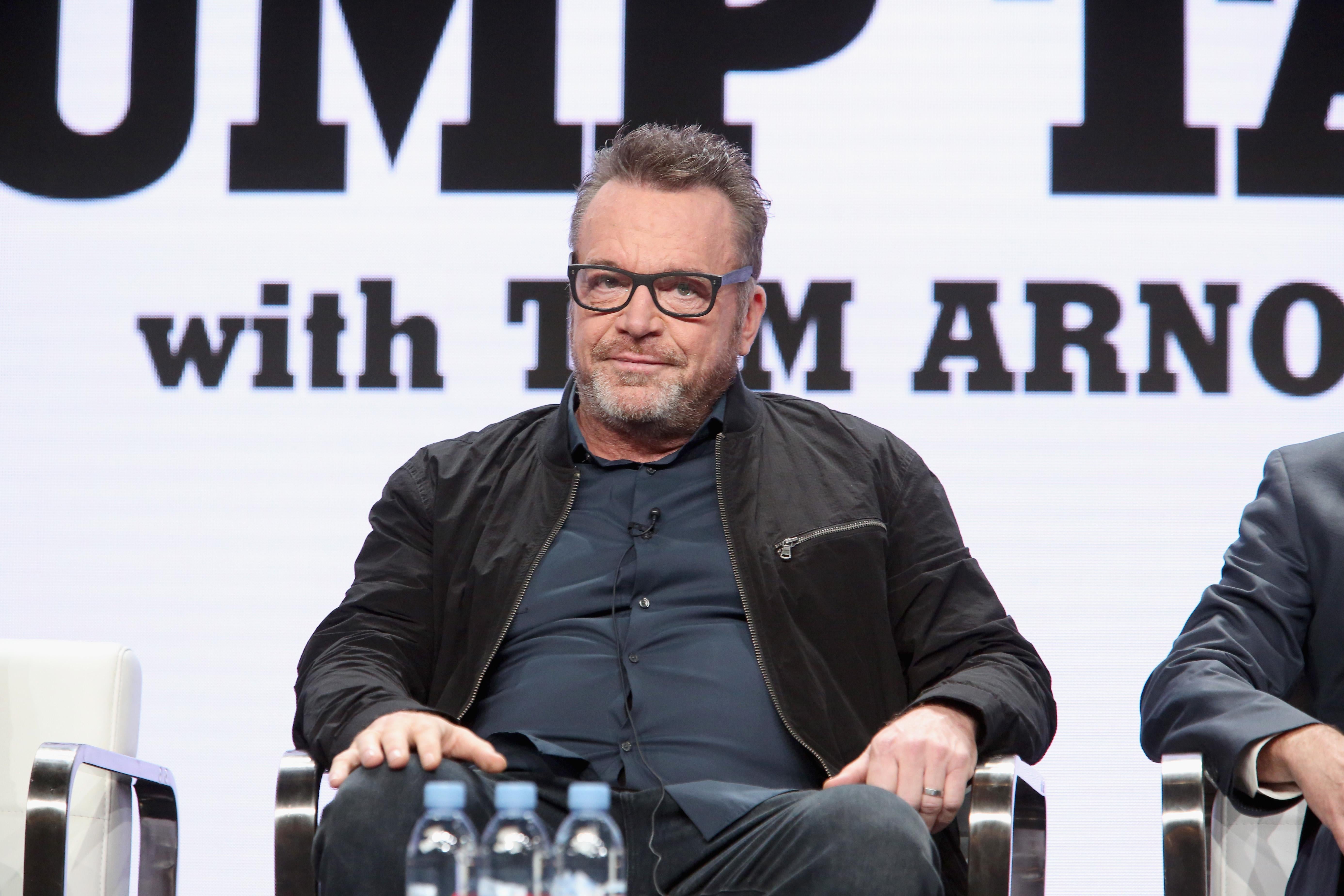 Tom Arnold sits at a panel on a black chair, wearing black-rimmed glasses and a black jacket