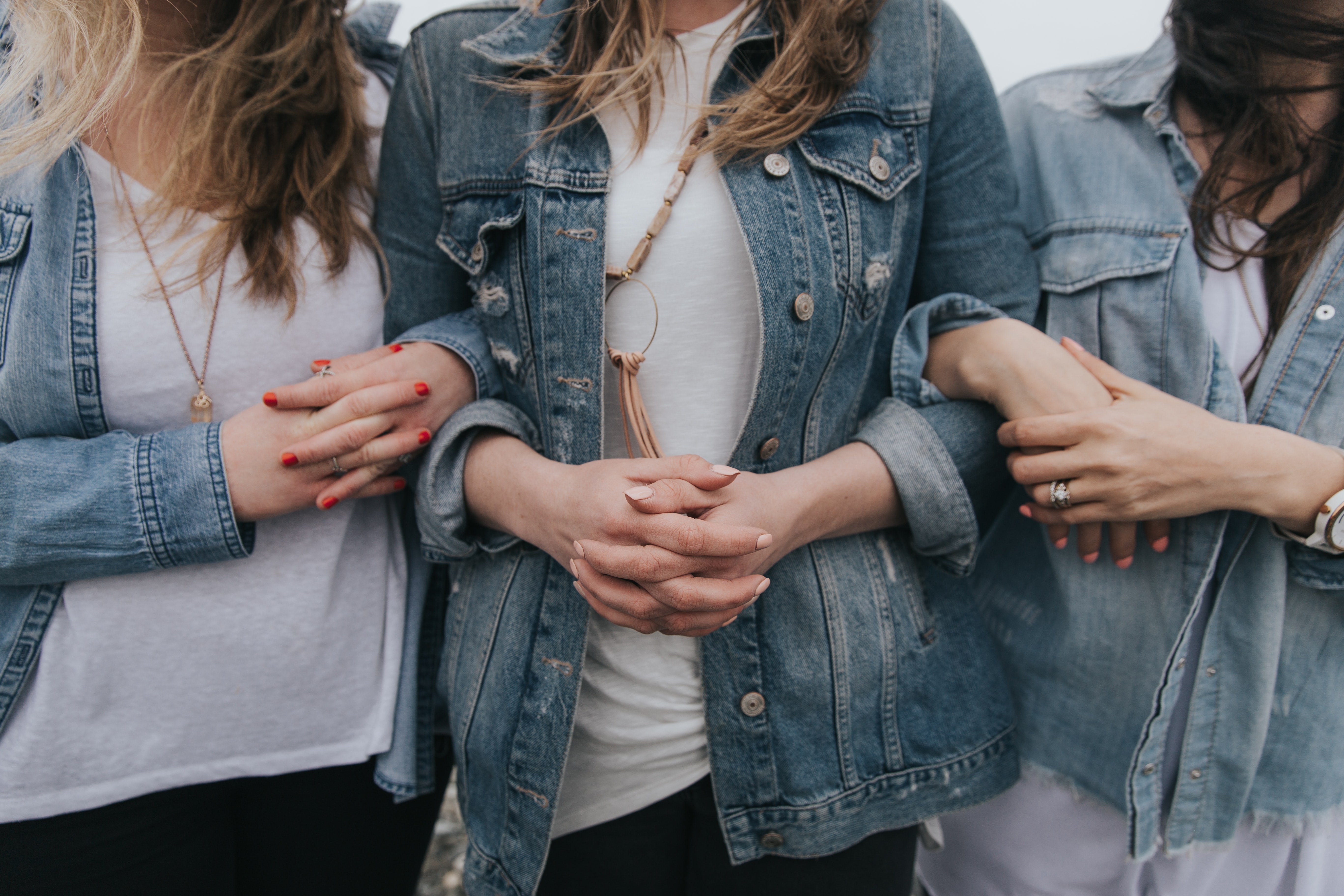 Three women photographed from the neck down, wearing white tees and oversized jean jackets. They are linking arms.