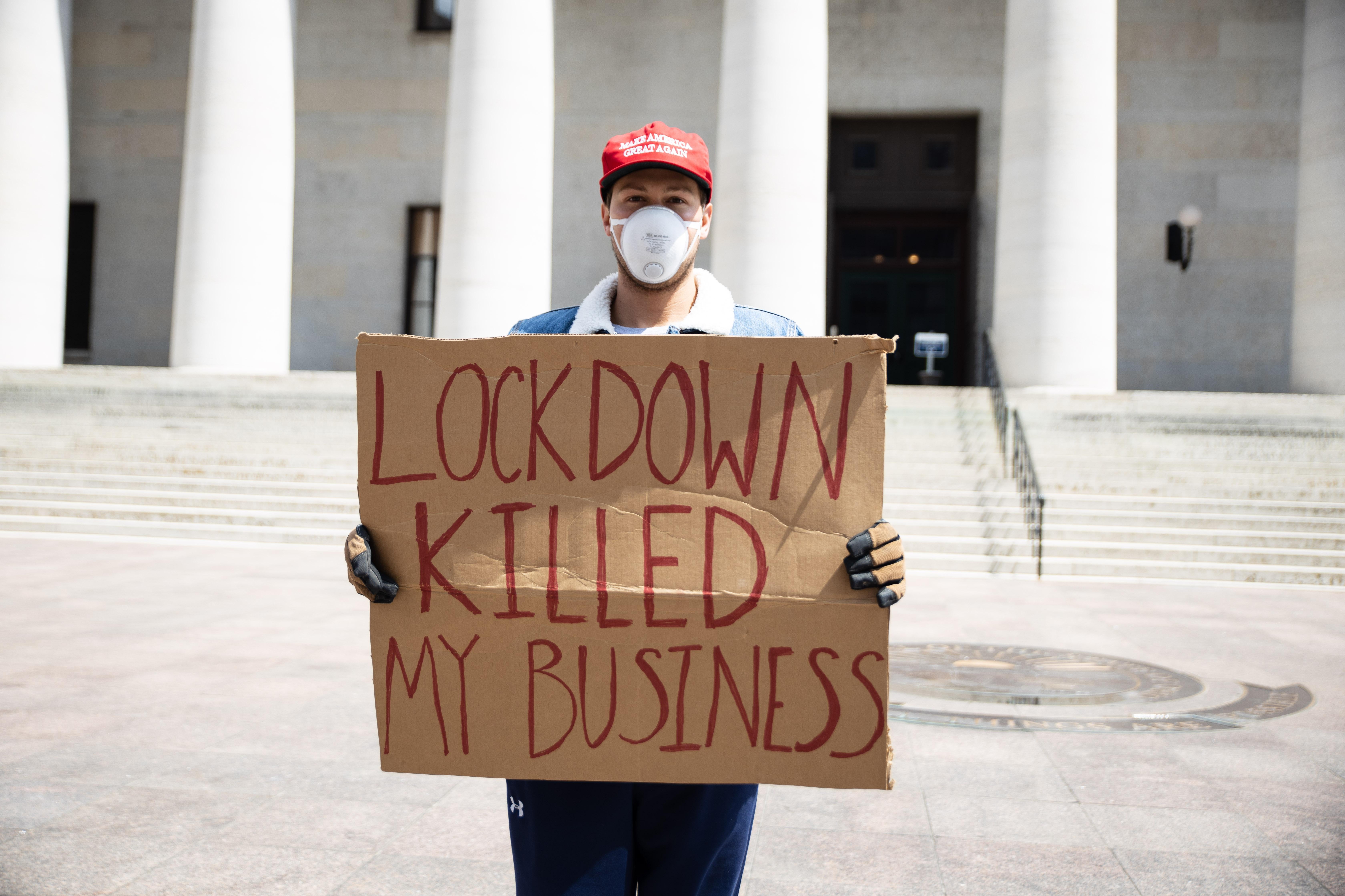 A protester wearing a MAGA hat and a medical mask holds a sign that says "Lockdown killed my business"